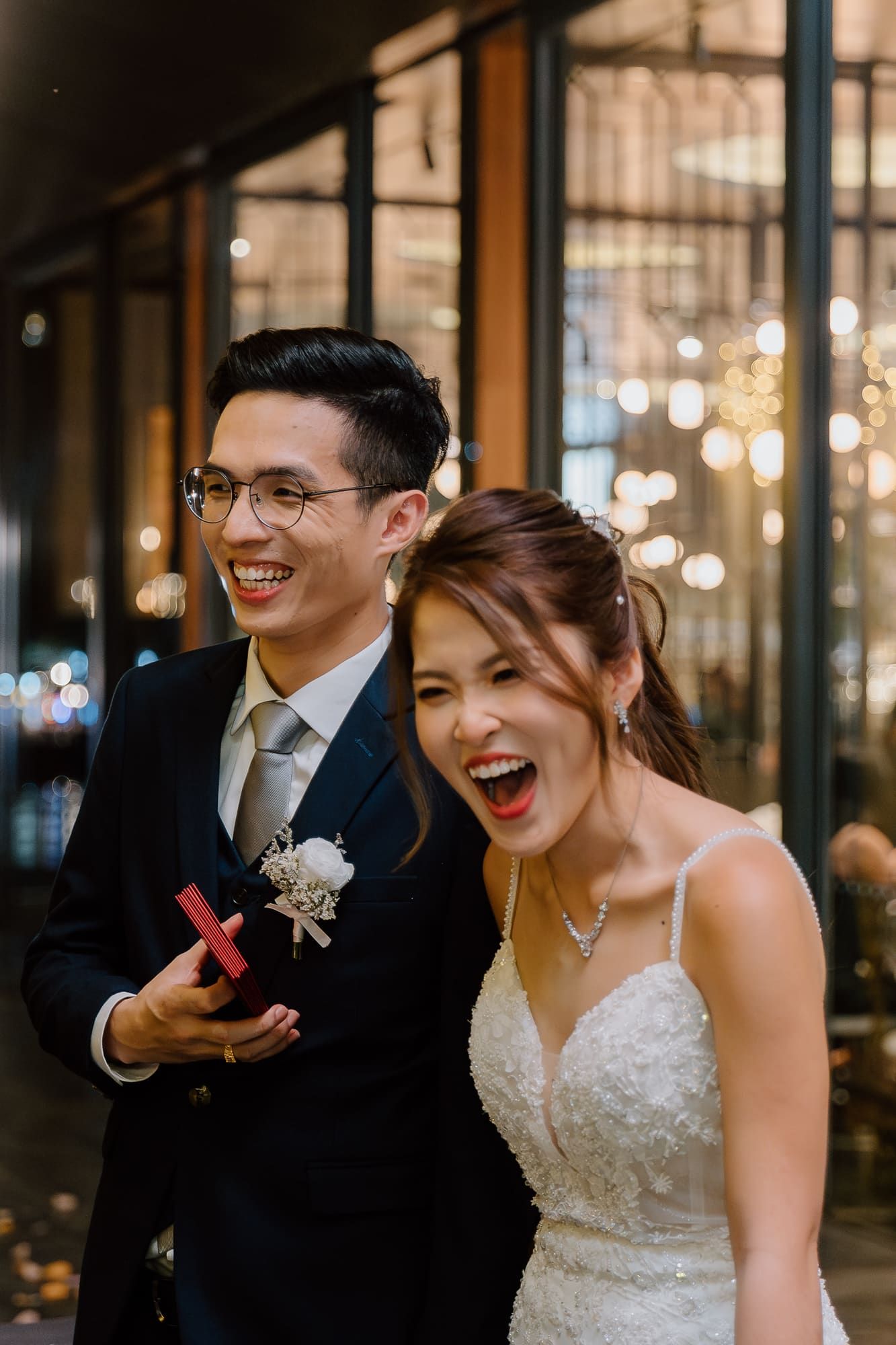 Couple shares joyful laughter during evening wedding celebration with glowing lights in background.