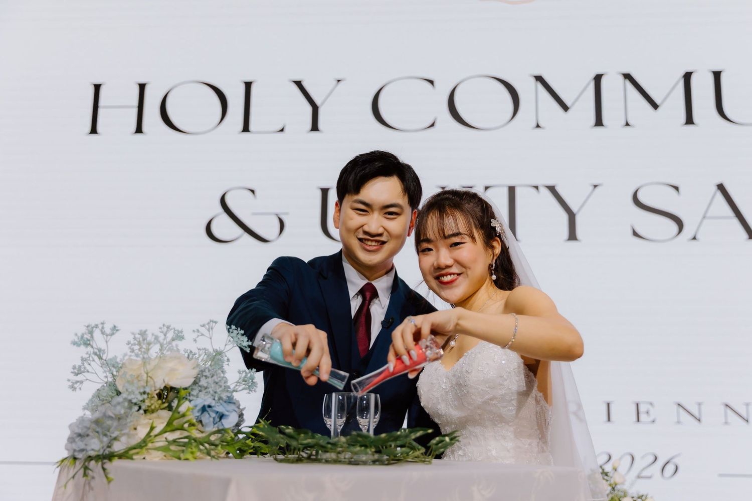 A smiling couple pours champagne together at their Holy Communion and unity ceremony wedding reception.