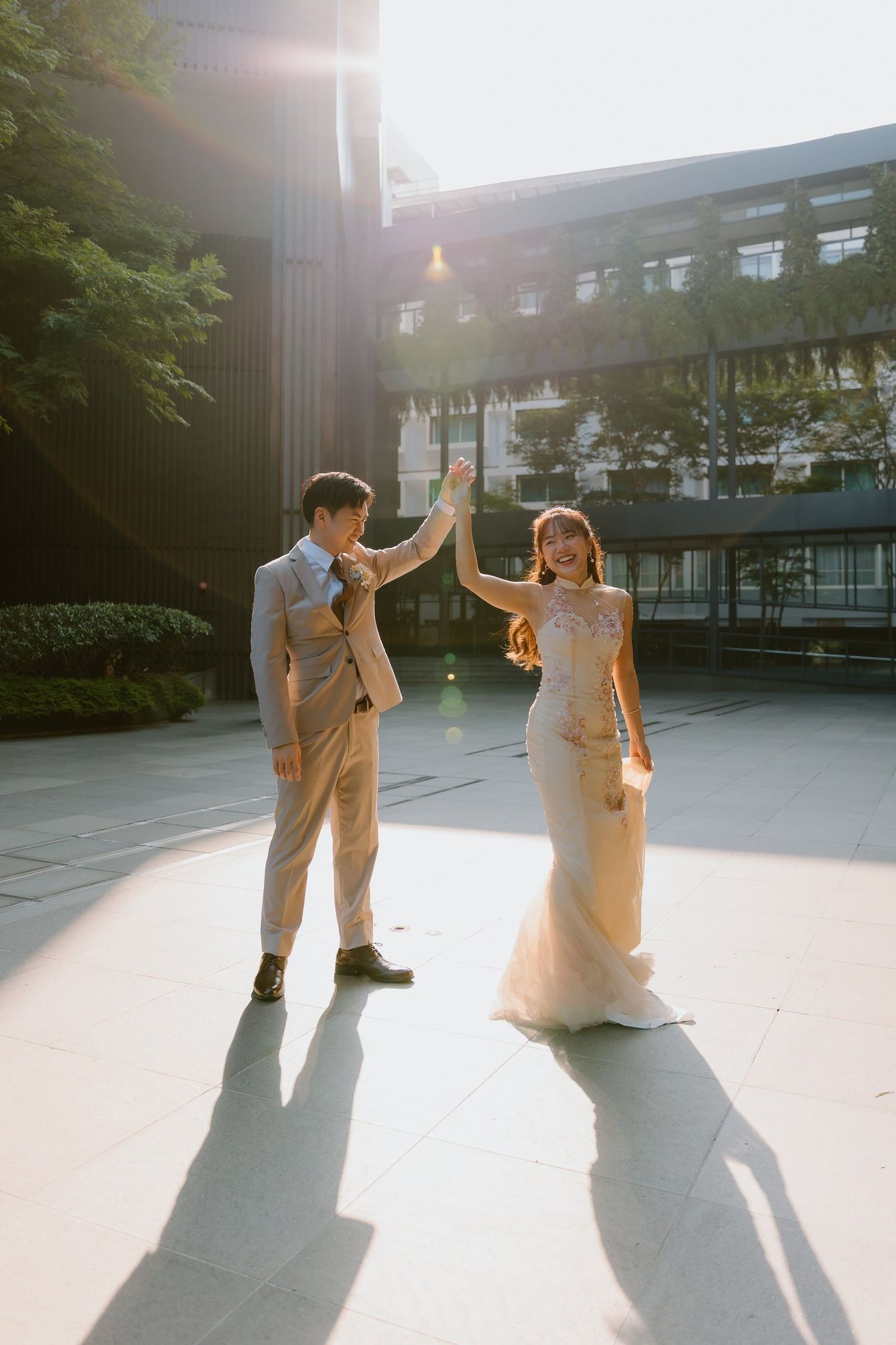 A couple in wedding attire holds hands and dances outdoors in golden sunlight, the bride in a mermaid gown and groom in a beige suit.