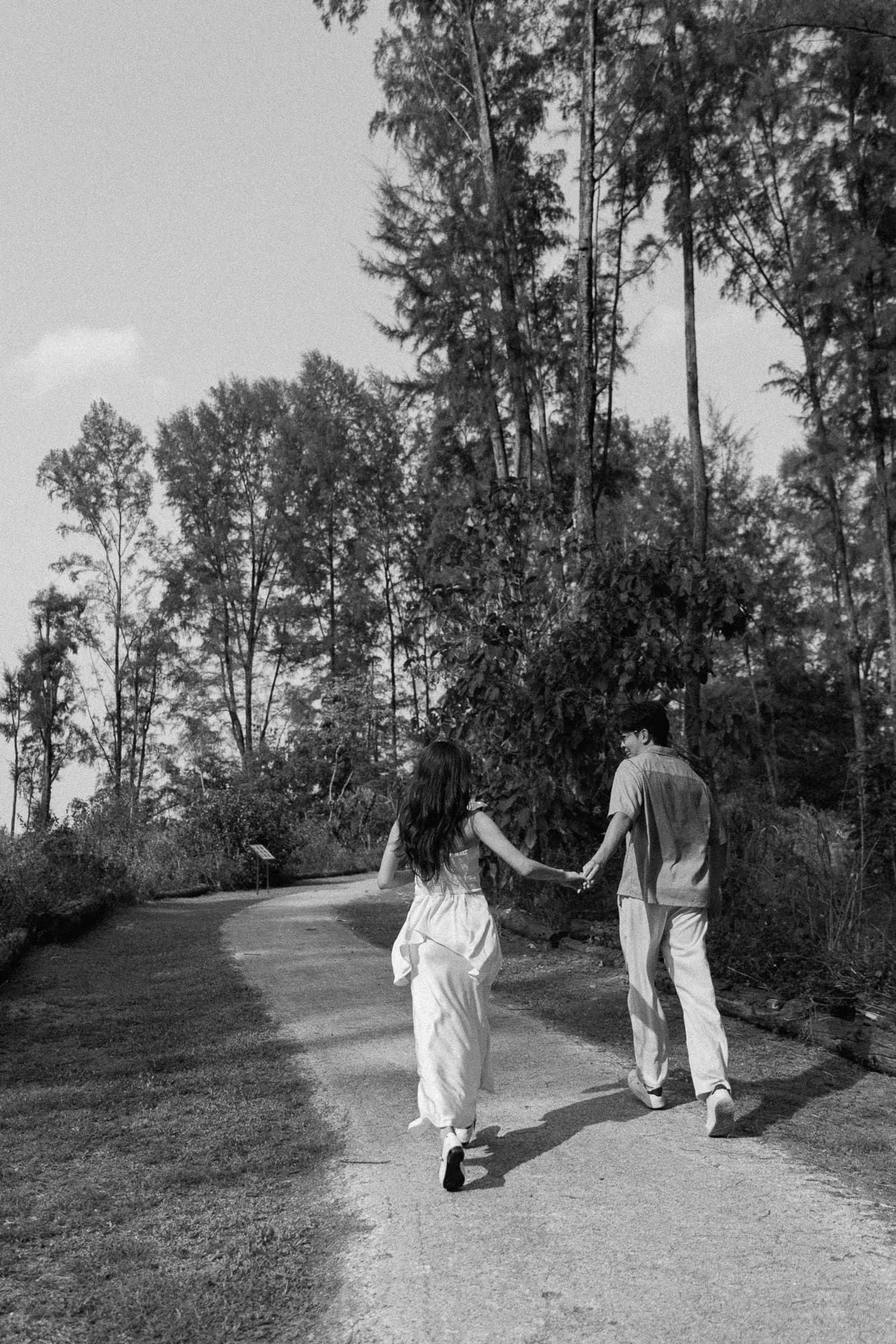 Black and white photo of a couple holding hands walking down a tree-lined path.