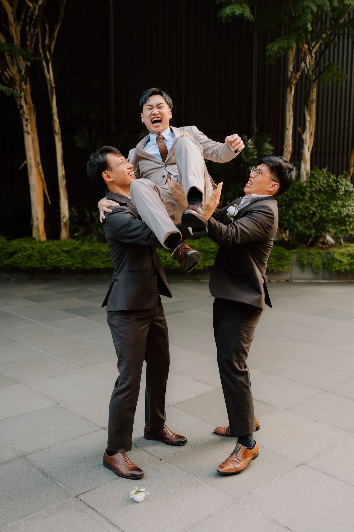 Two groomsmen in dark suits joyfully lift a laughing groom in a tan suit outdoors at a wedding celebration.