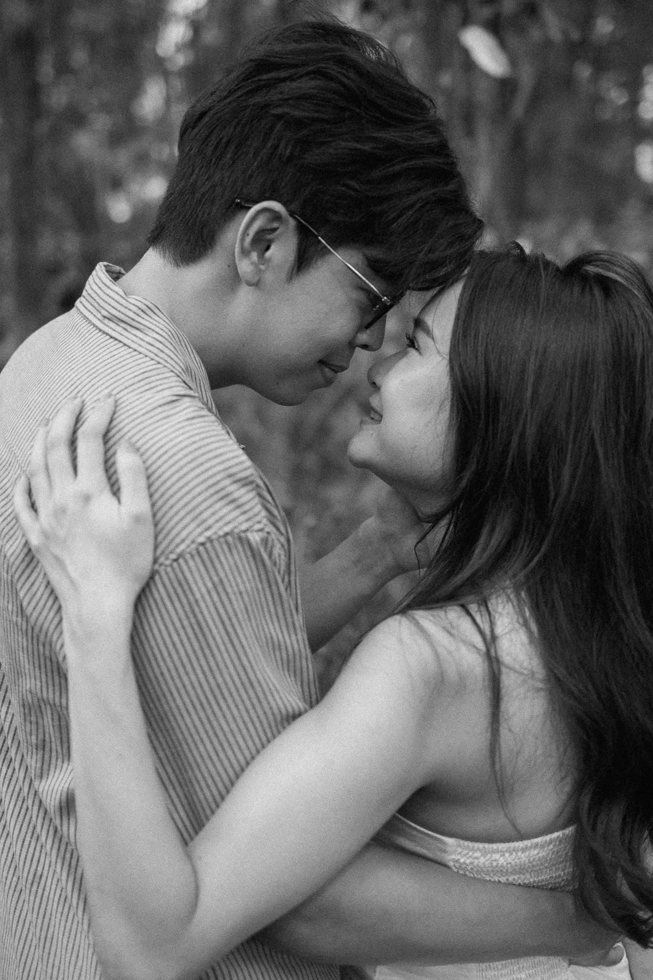 Black and white photo of a young couple embracing and about to kiss outdoors among trees.