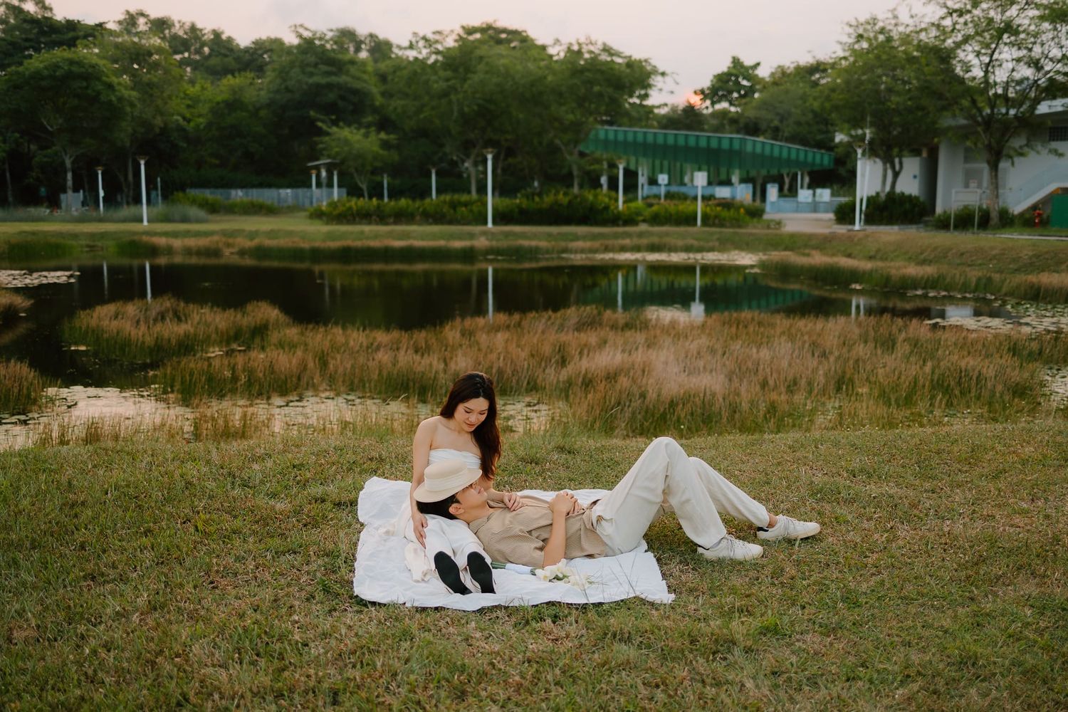 Couple relaxing on white blanket by pond in lush green park during golden hour.