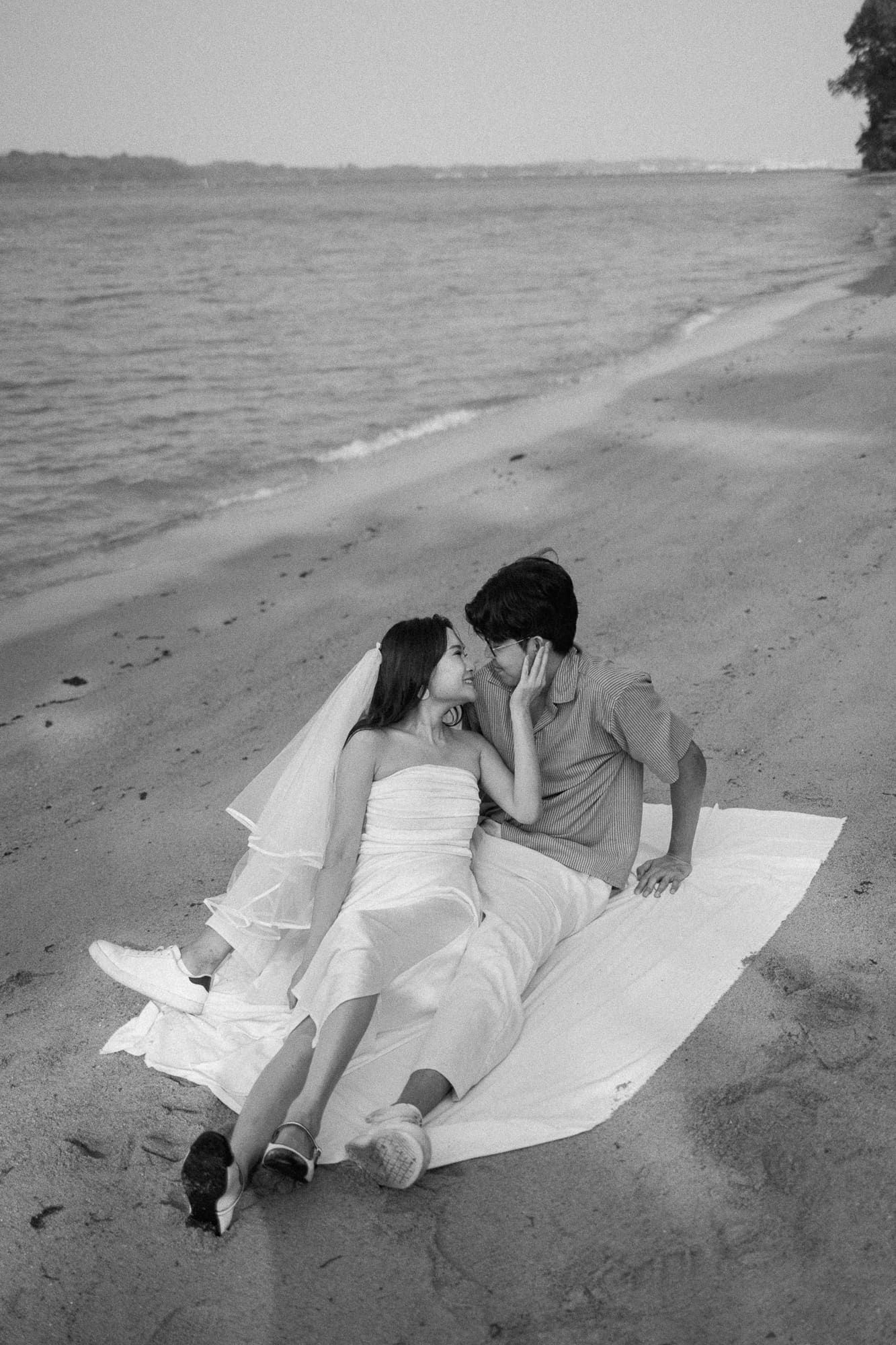 Black and white photo of a bride and groom sitting together on a sandy beach, sharing a romantic moment by the ocean.