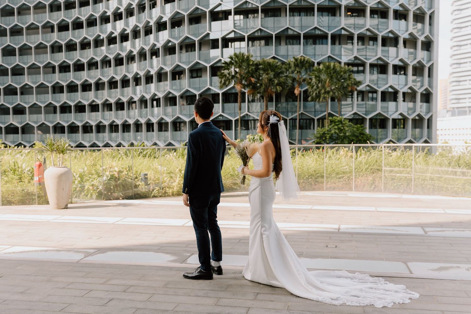 Elegant couple in formal attire stands in front of a modern architectural building with honeycomb-patterned facade and palm trees.