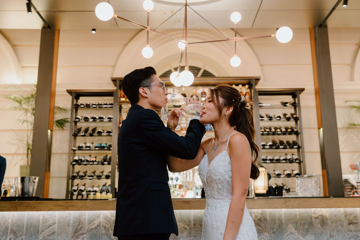 A couple shares a loving moment at a wine bar counter with illuminated pendant lights and wine racks in the background.
