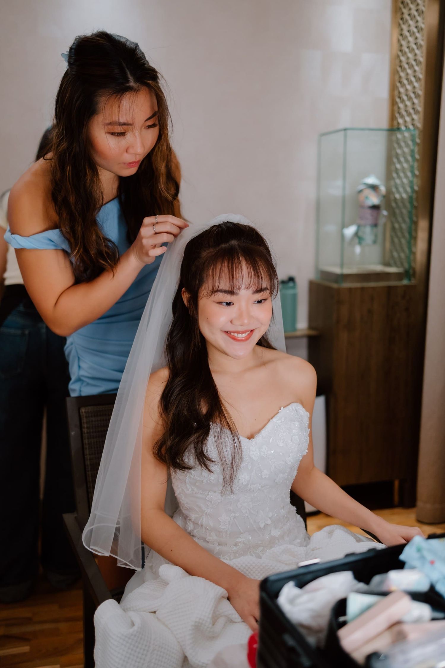A bride in a white gown smiles as her bridesmaid adjusts her veil during wedding preparations.