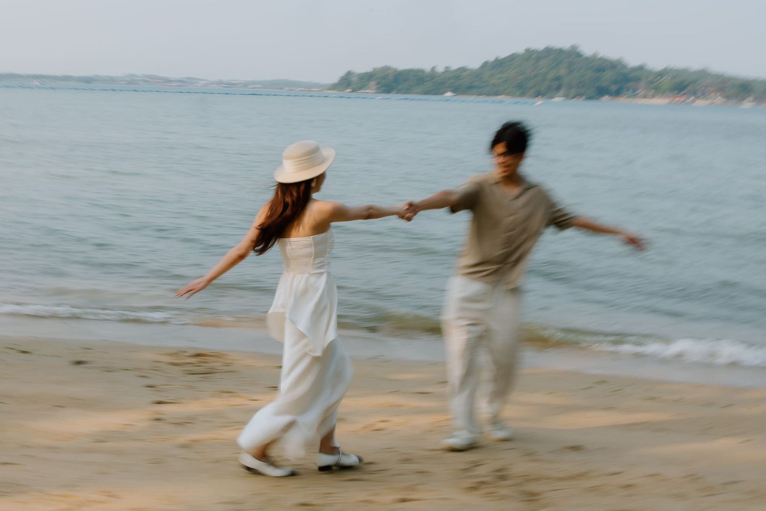 Couple holding hands dancing playfully on a sandy beach with calm ocean waves and green island in background.