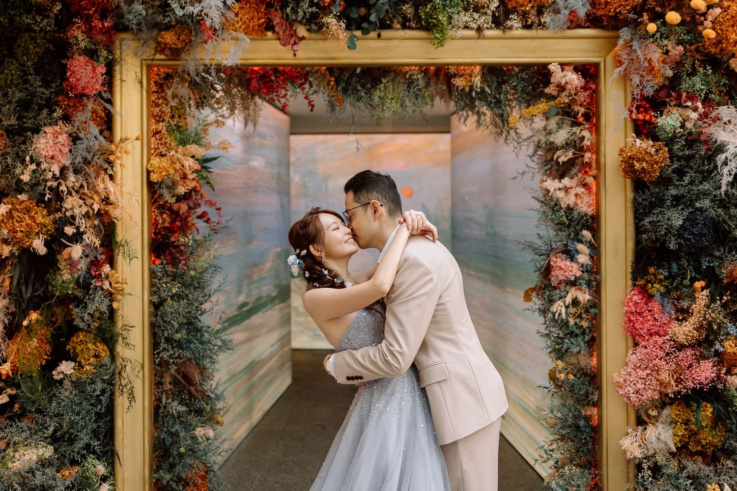 Couple kissing under ornate floral arch with autumn blooms at wedding.