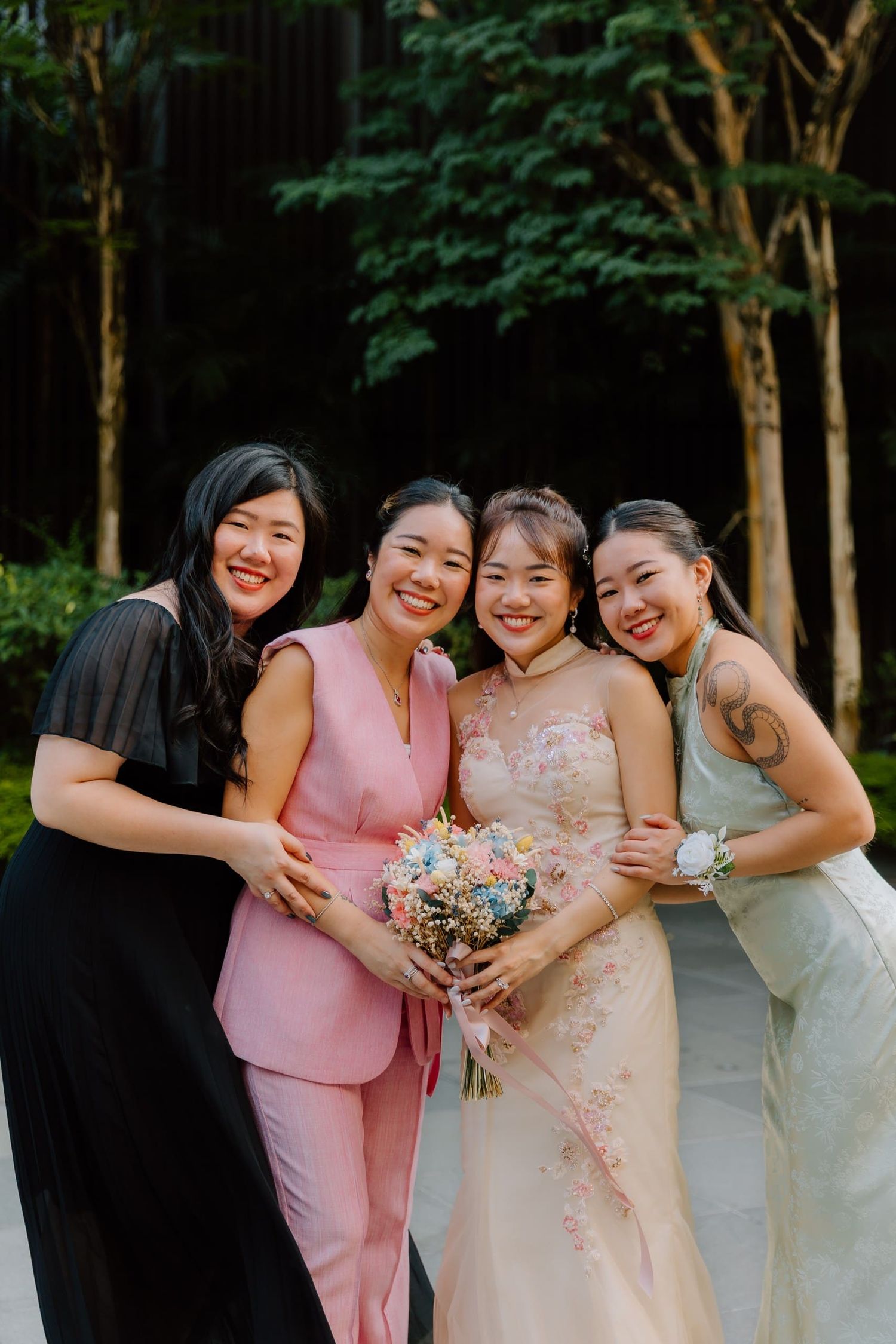 Bride in floral gown holds bouquet surrounded by three smiling bridesmaids in black, pink, and sage green outdoors.