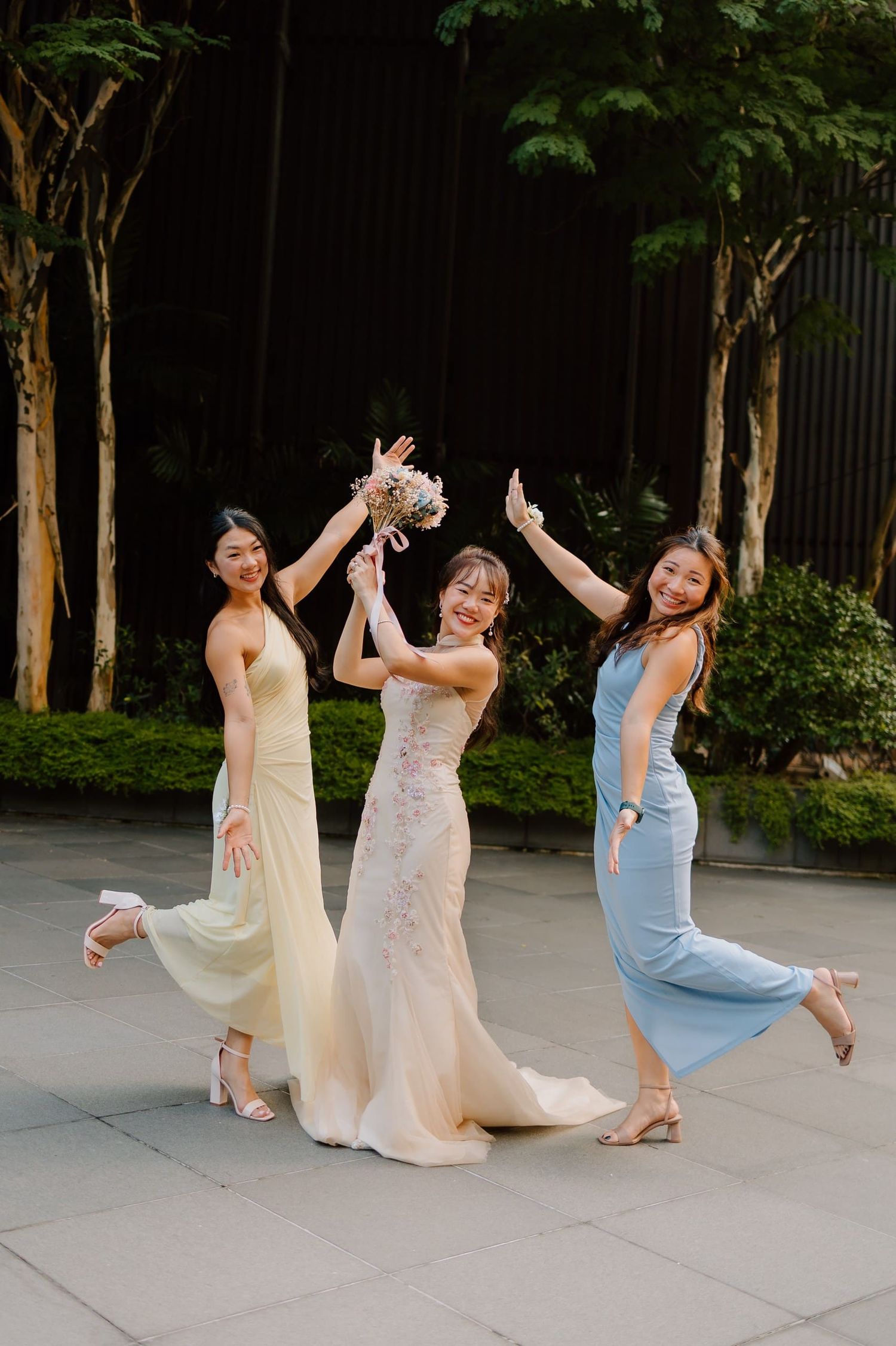 Bride in white gown celebrates with two bridesmaids in yellow and blue dresses, holding bouquet outdoors.
