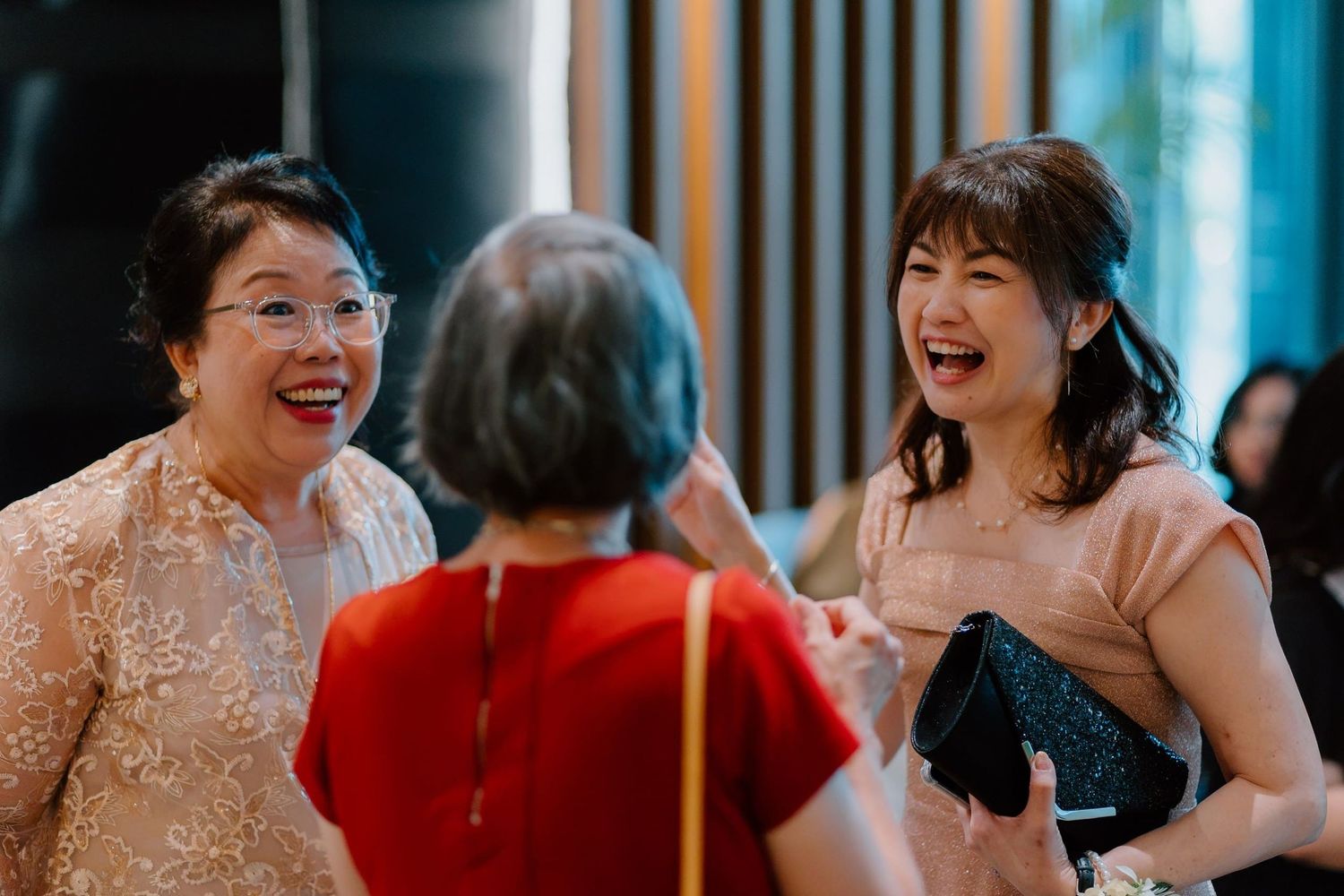 Three Asian women laughing and socializing joyfully at a formal event or wedding celebration.