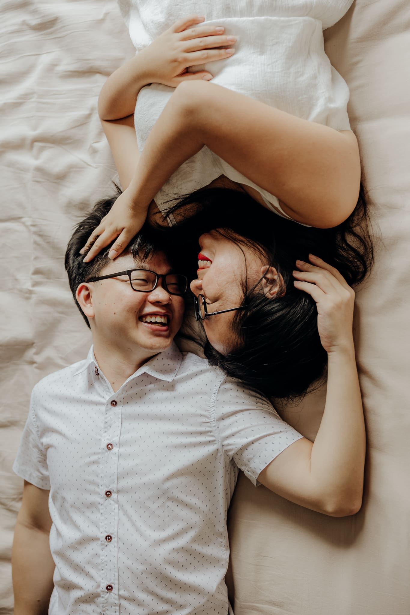 Couple sharing laughter and tender moments while lying on white sheets.