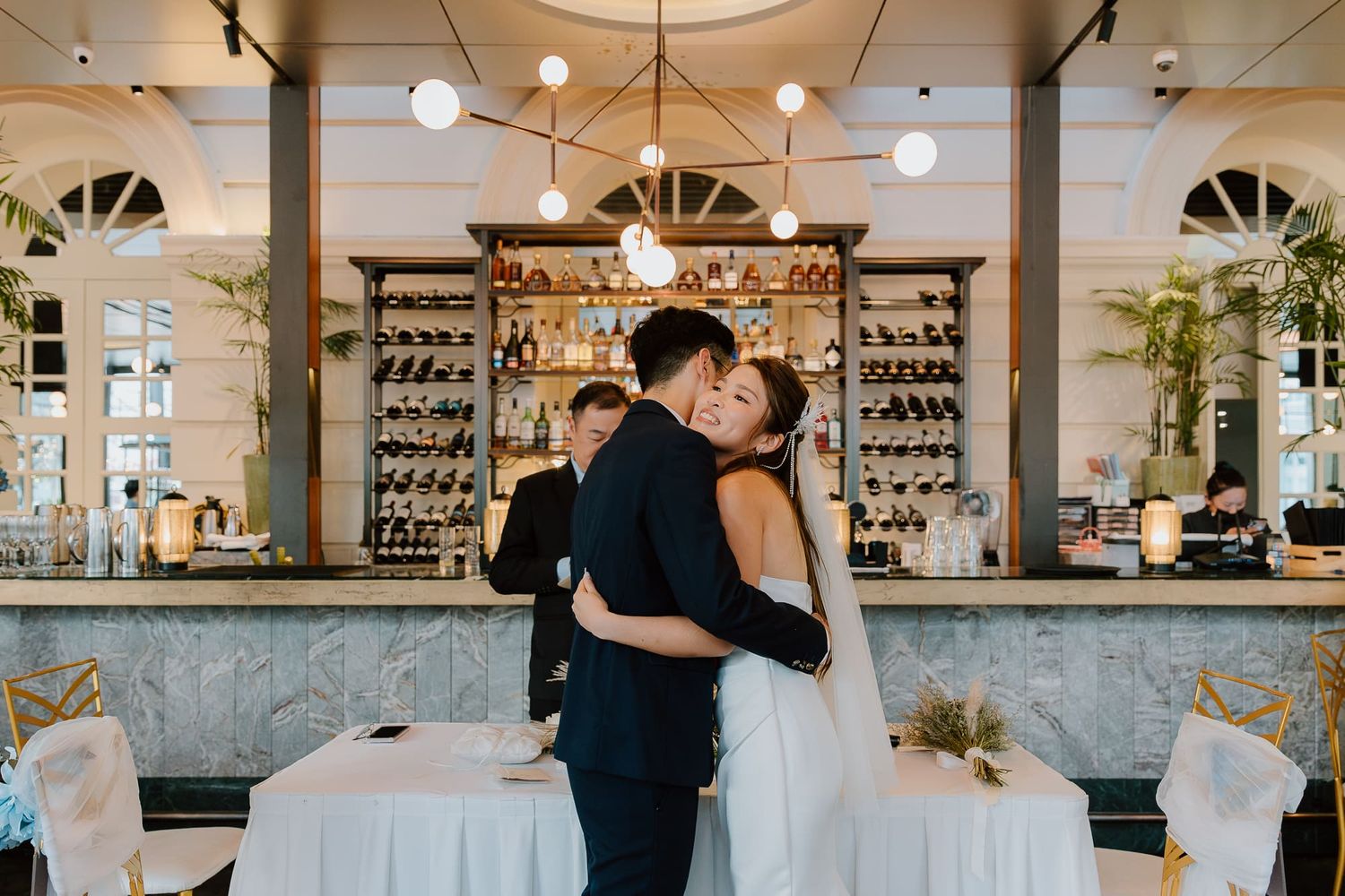 Newlyweds embrace on their wedding day in an elegant restaurant with a vintage bar and glowing pendant lights.