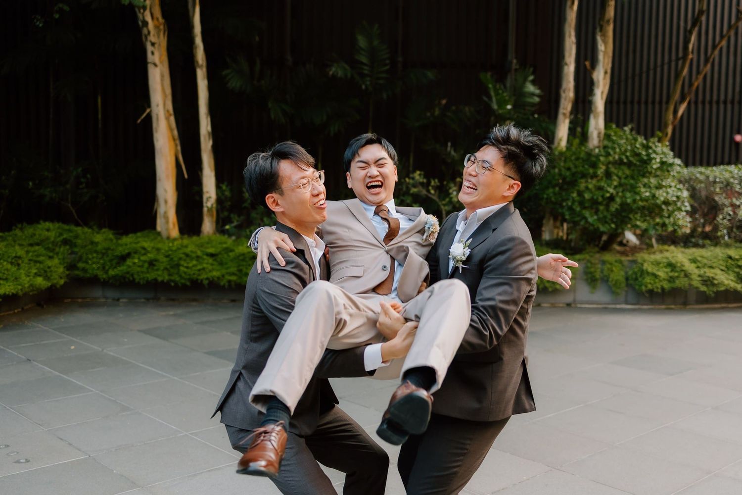 Three groomsmen laughing joyfully outdoors, two lifting the groom in a celebratory wedding photo.
