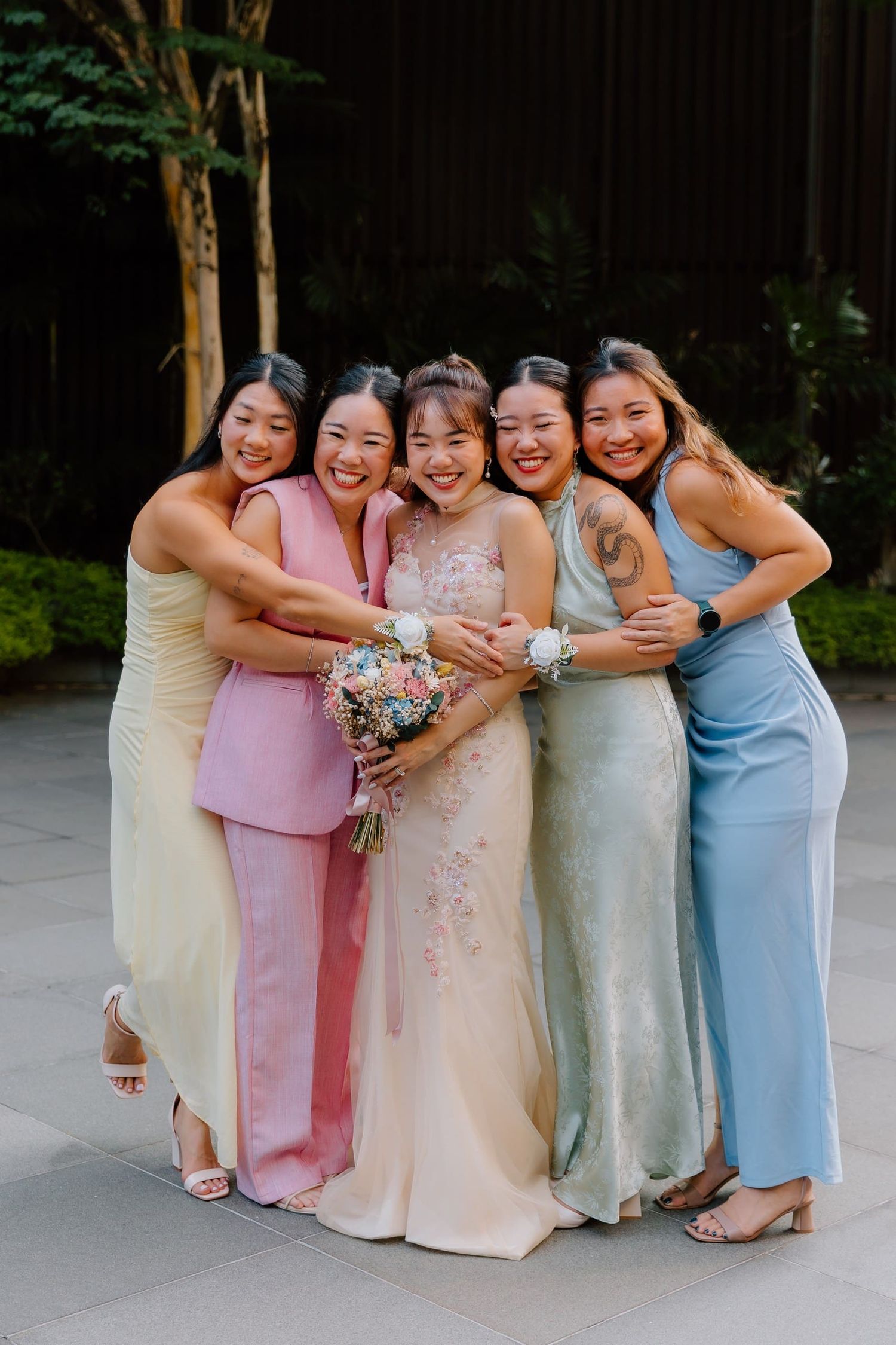 Bride in embroidered gown hugged by five bridesmaids in colorful dresses, smiling together at outdoor wedding venue.