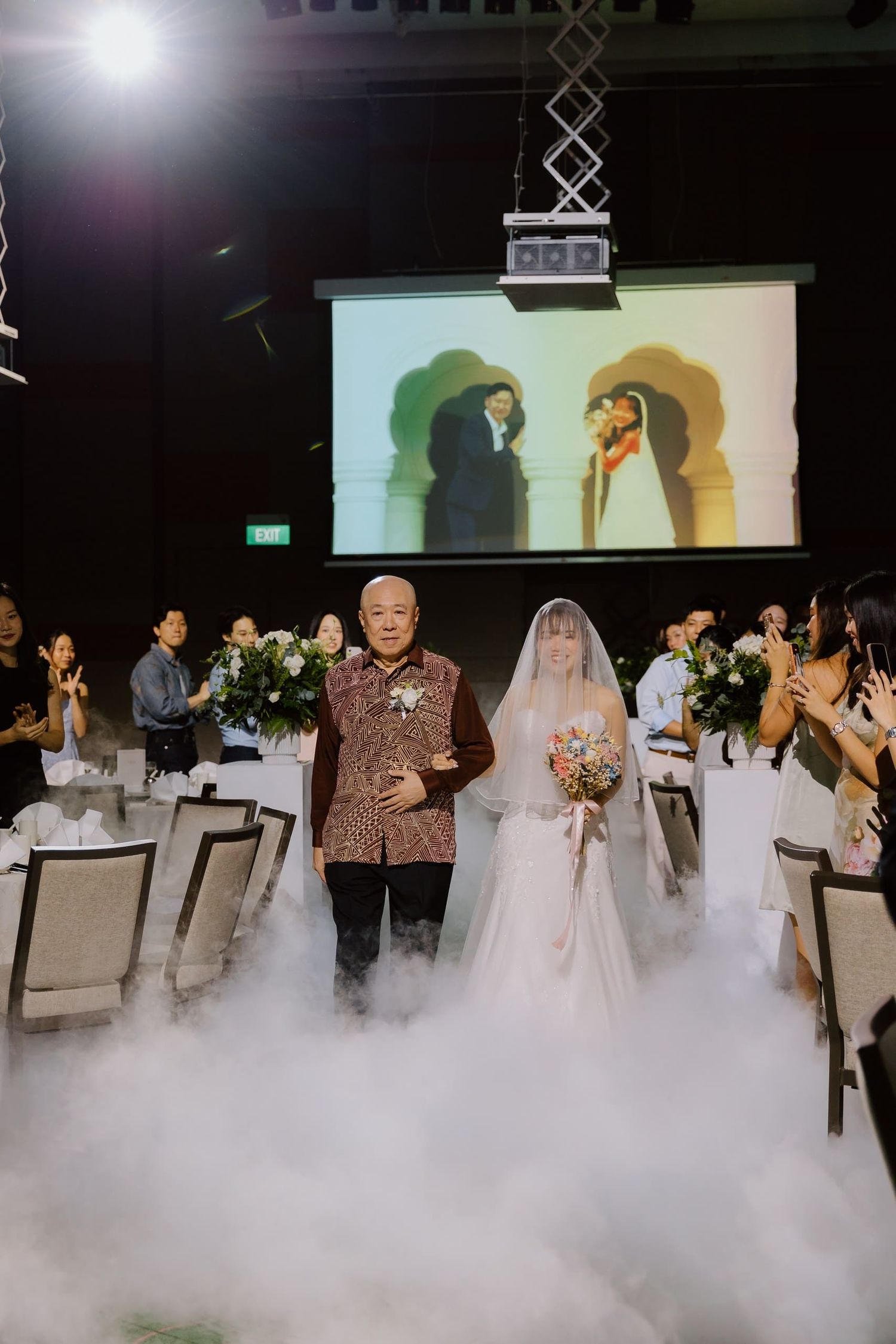 Bride walks down the aisle with her father through dramatic fog at an elegant wedding reception venue.