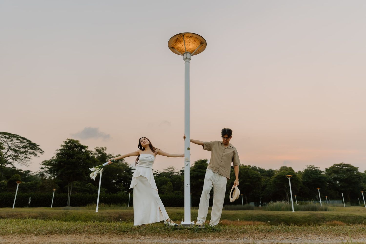 Couple in white outfits posing playfully by a lamp post at sunset in a lush green park.