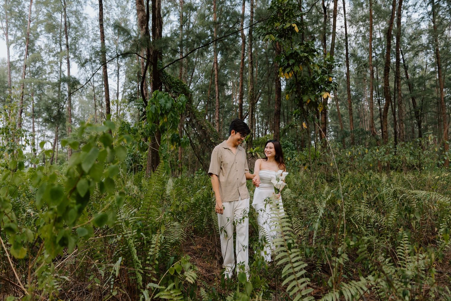 Couple in neutral outfits walking through lush forest ferns, bride holding bouquet, romantic outdoor pre-wedding photoshoot.