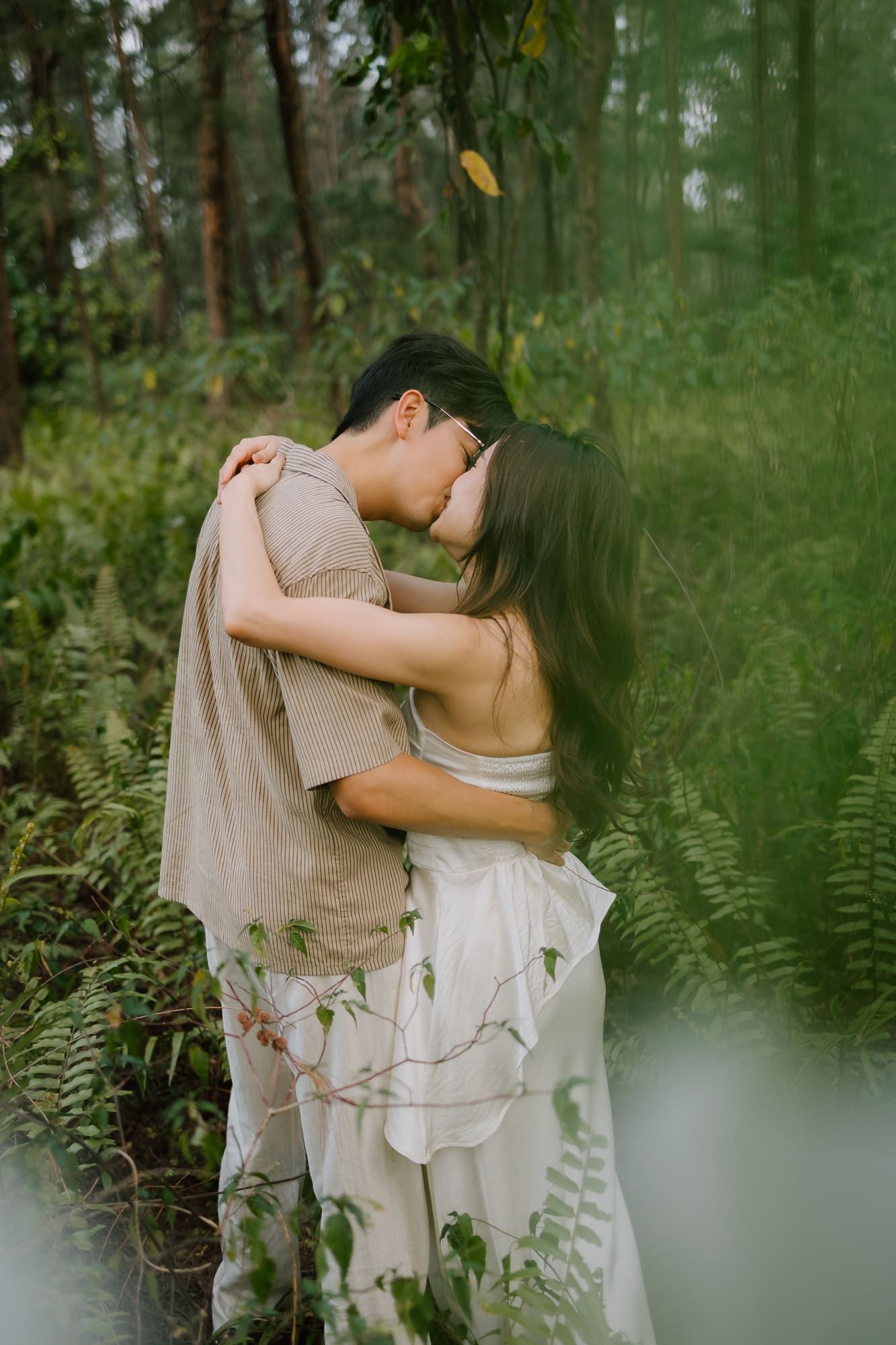 A couple shares a romantic kiss while embracing in a lush green forest, wearing neutral linen and white dress.