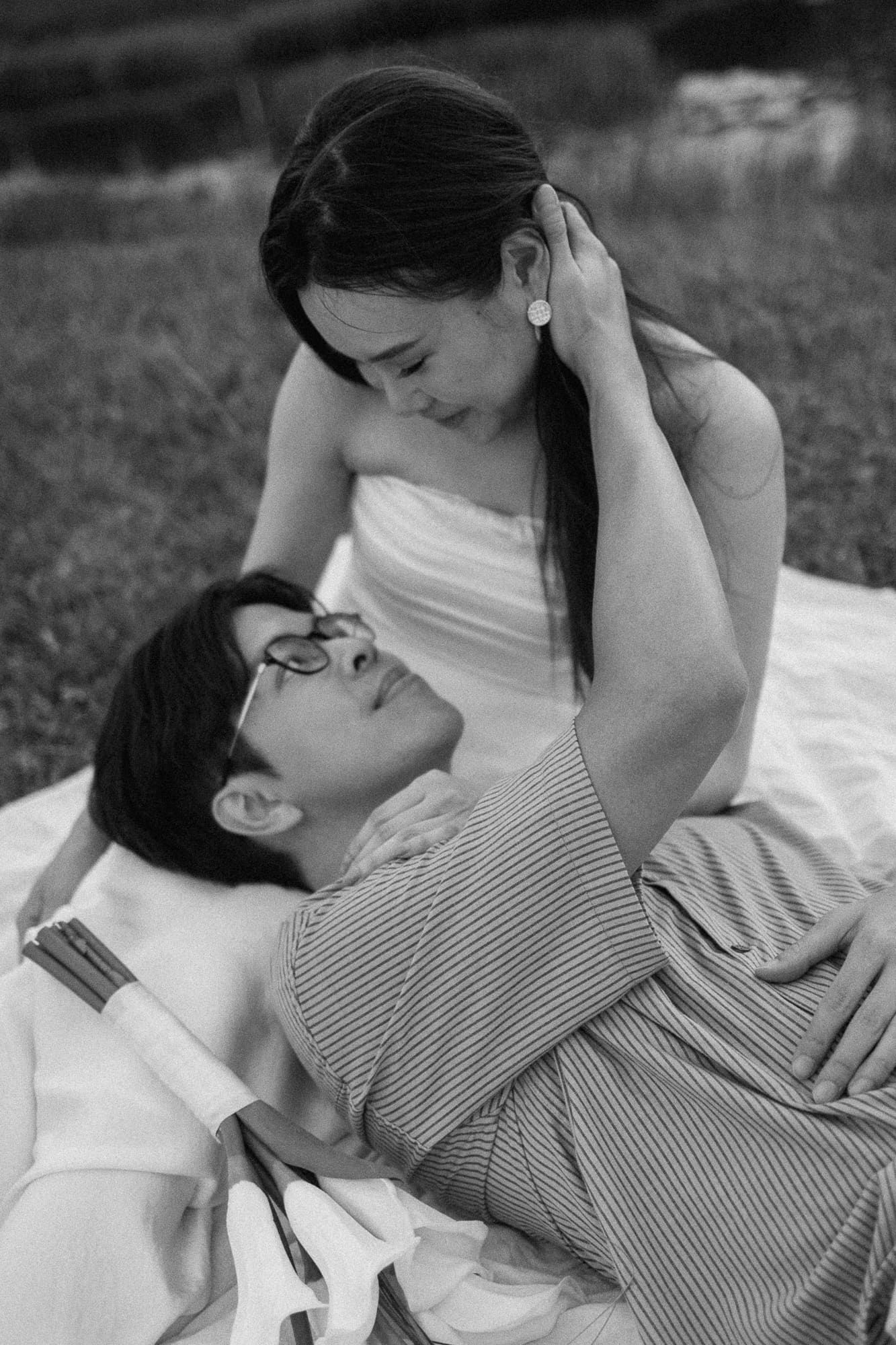 Black and white photo of a couple sharing a tender moment on a blanket outdoors, woman in white dress gazing at man.