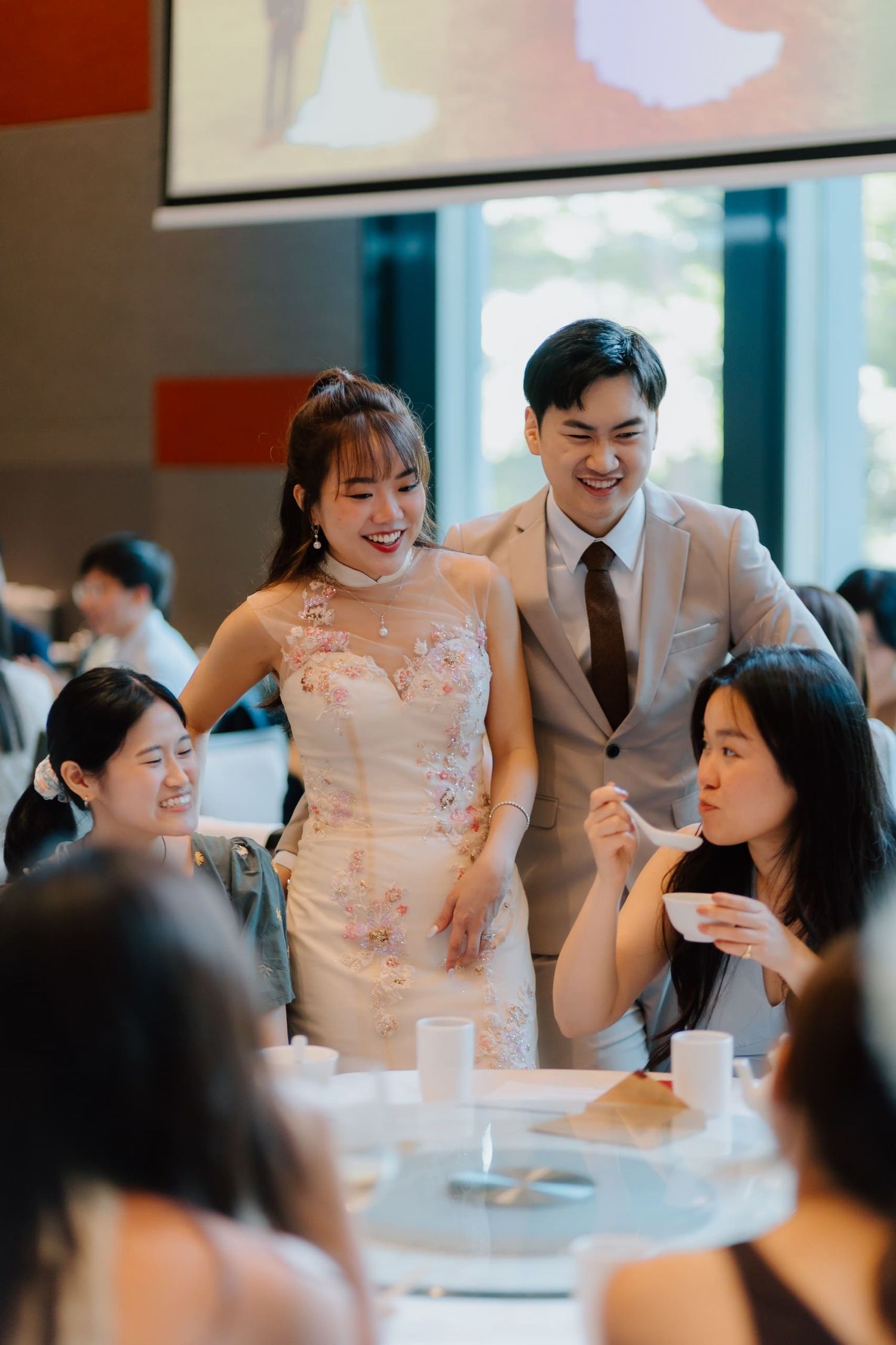 Smiling bride in floral gown and groom in suit greeting guests at wedding reception.