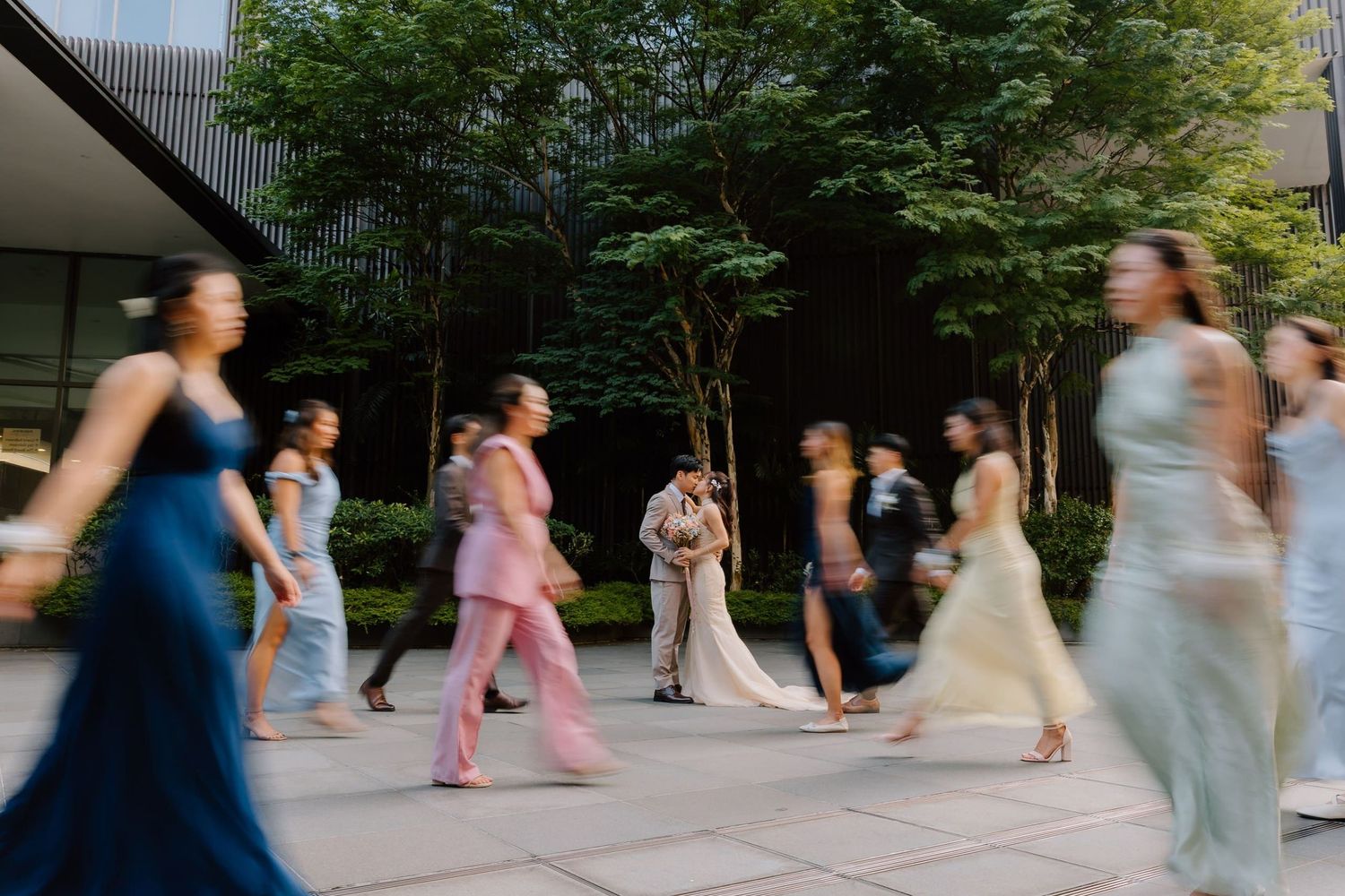 Blurred motion shot of wedding party in colorful gowns walking outdoors near trees, with bride and groom in center.