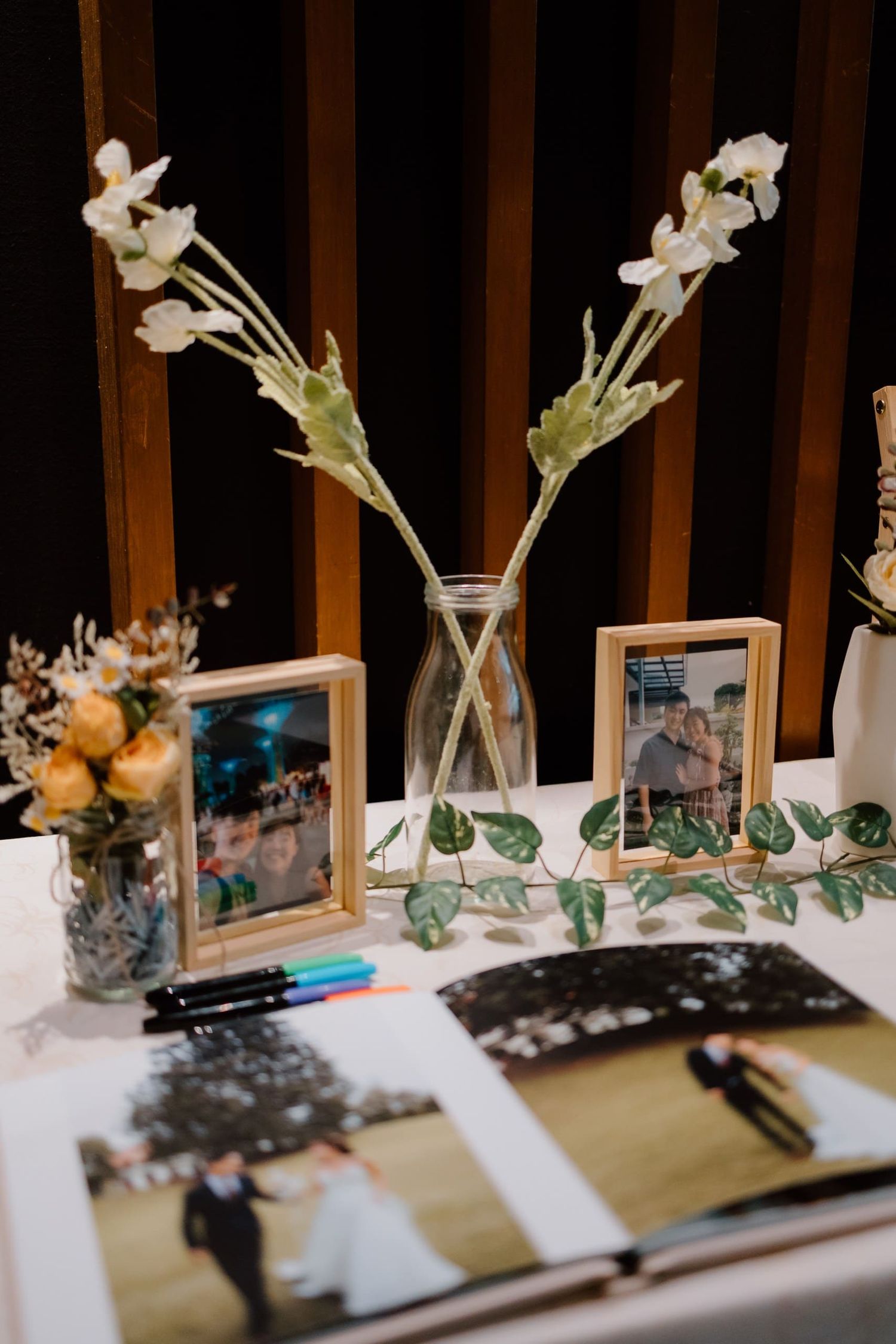 Wedding guest book table with framed couple photos, white floral stems in glass vase, markers, and open photo album.