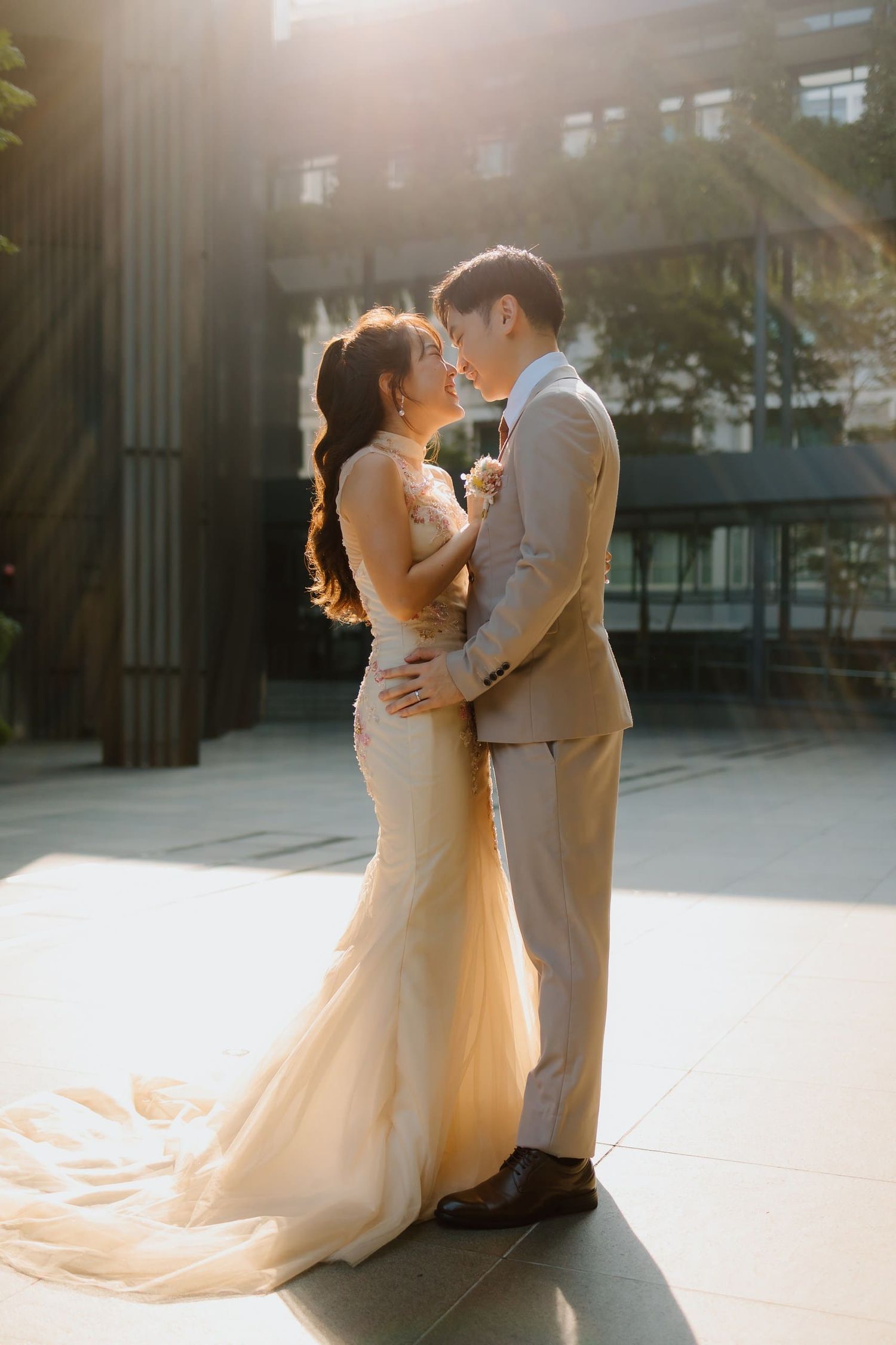 Bride in elegant gown and groom in tan suit embrace in golden sunset light outdoors.