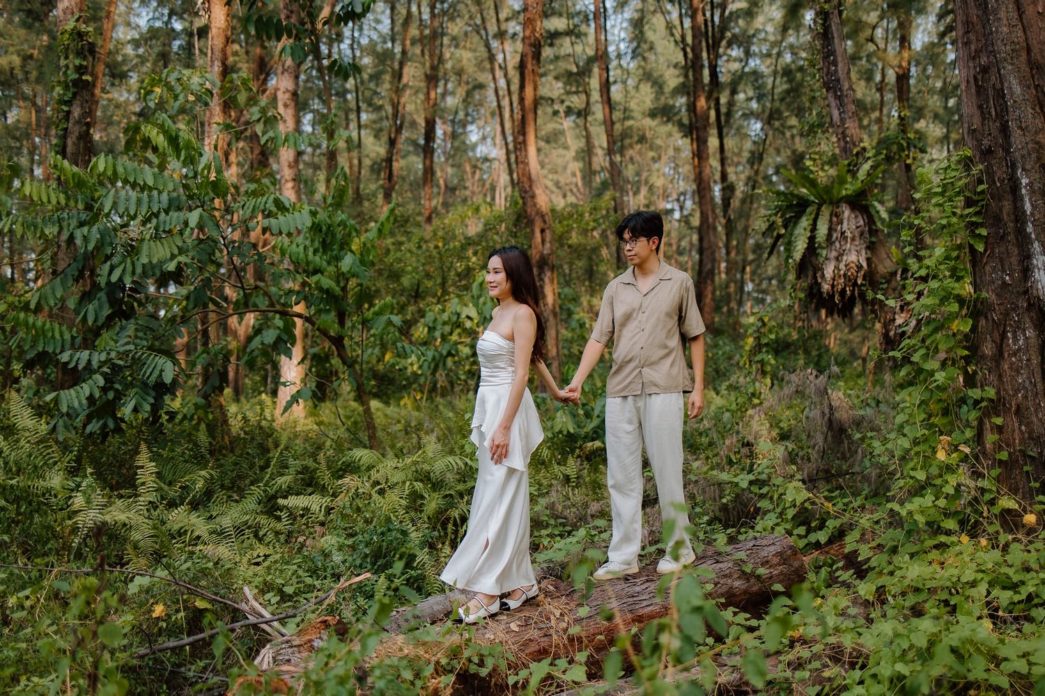 Couple holding hands in a lush forest, woman in white dress, man in beige outfit, standing on a log.