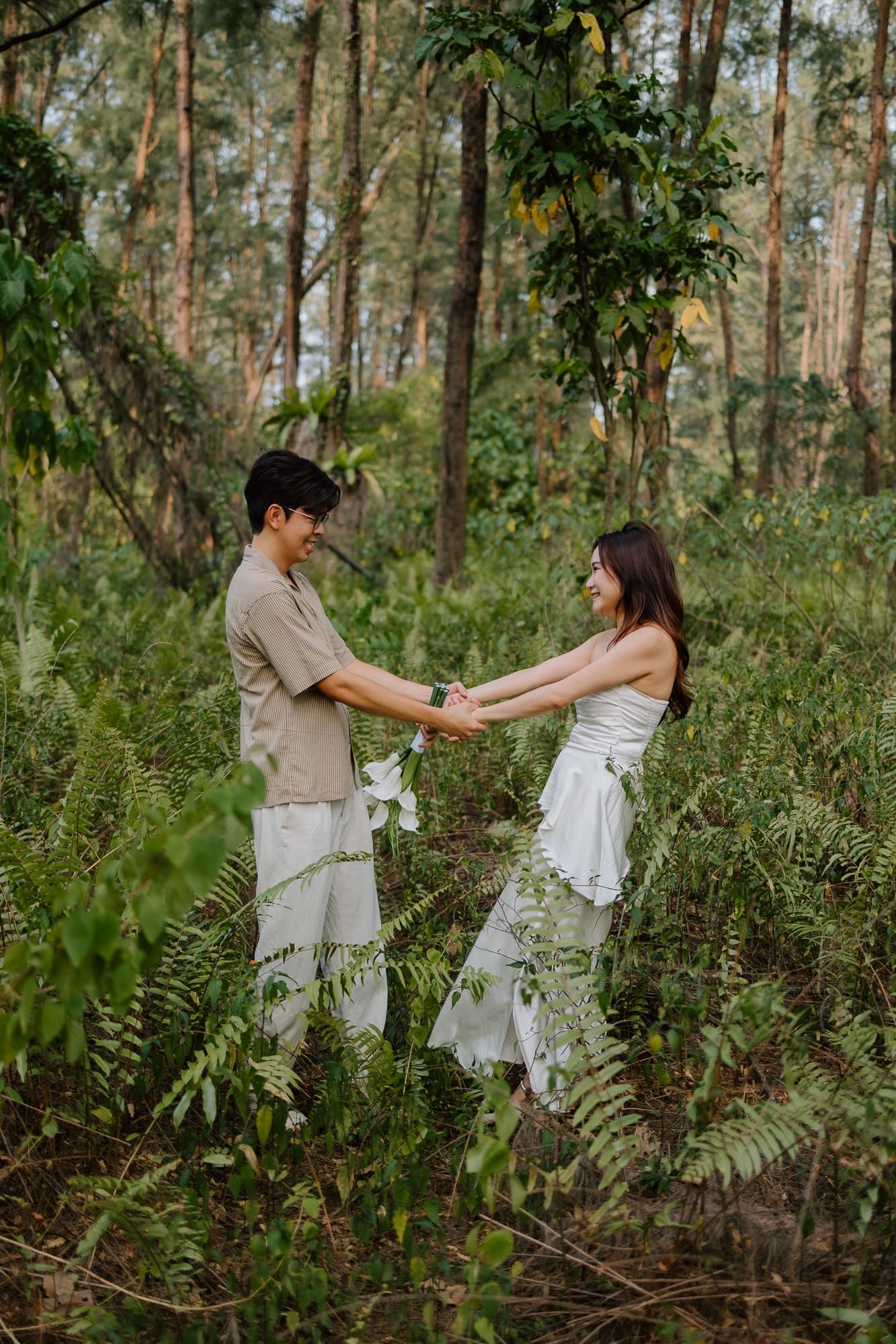 A couple holds hands and smiles at each other in a lush green forest, man in beige outfit, woman in white dress.