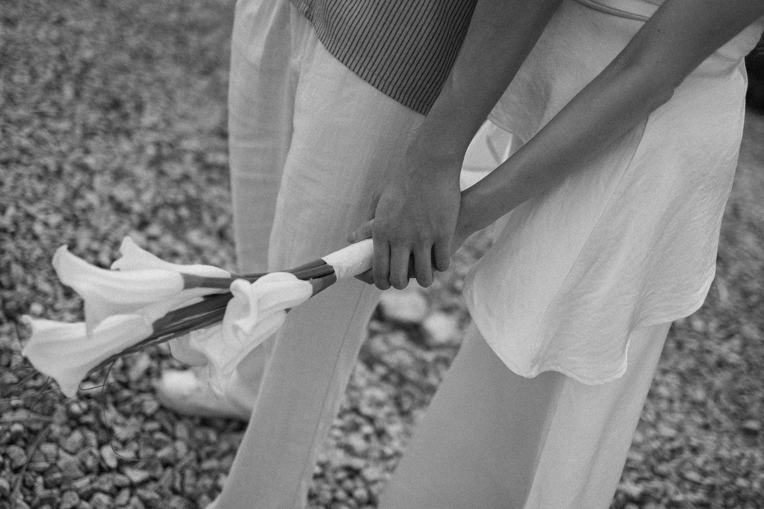 Black and white photo of a ballerina adjusting her pointe shoes while holding a bouquet near a gravel path.