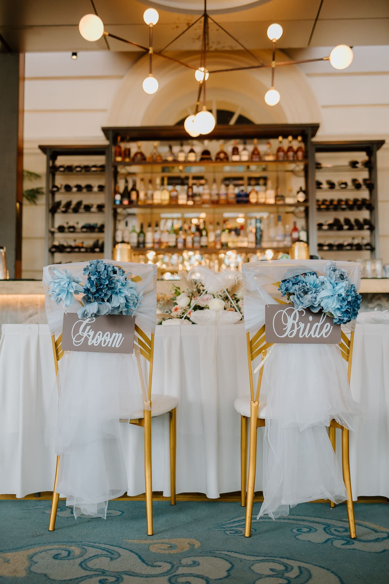 Elegant wedding reception chairs with 'Bride' and 'Groom' signs and blue hydrangea flowers at a bar venue.