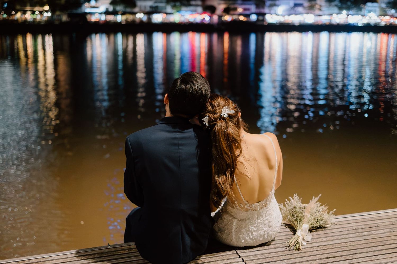 A couple sits on a wooden dock at night with city lights reflecting on the water as they share an intimate moment.