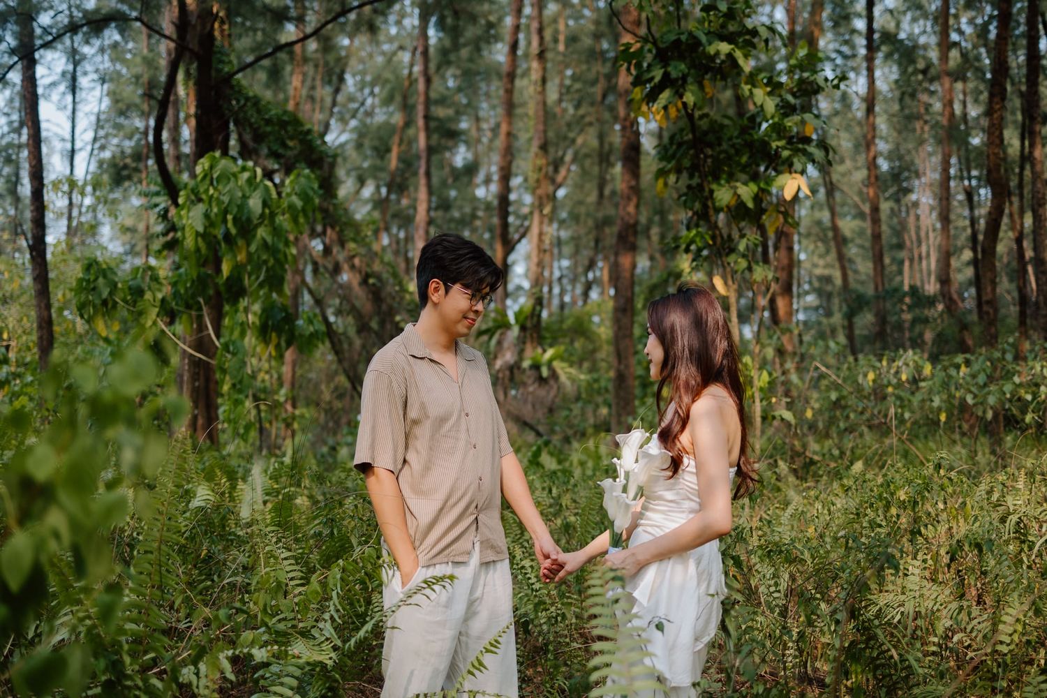 A couple holds hands in a lush forest, smiling at each other in casual outfits.