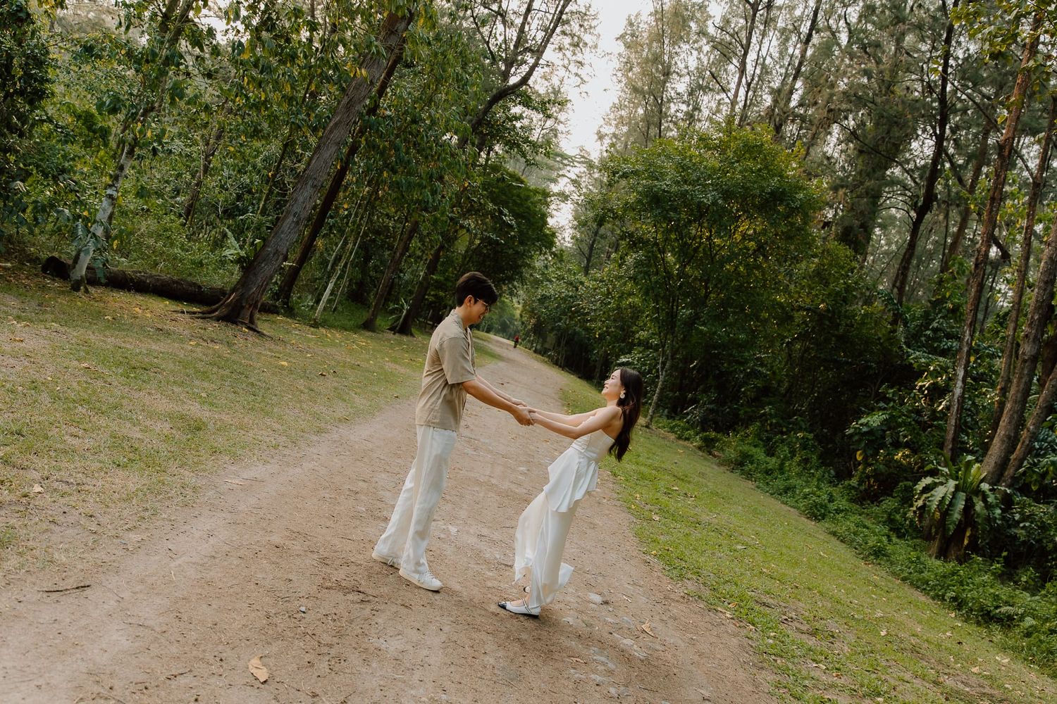 Couple holding hands and spinning on a forest dirt path surrounded by lush green trees.