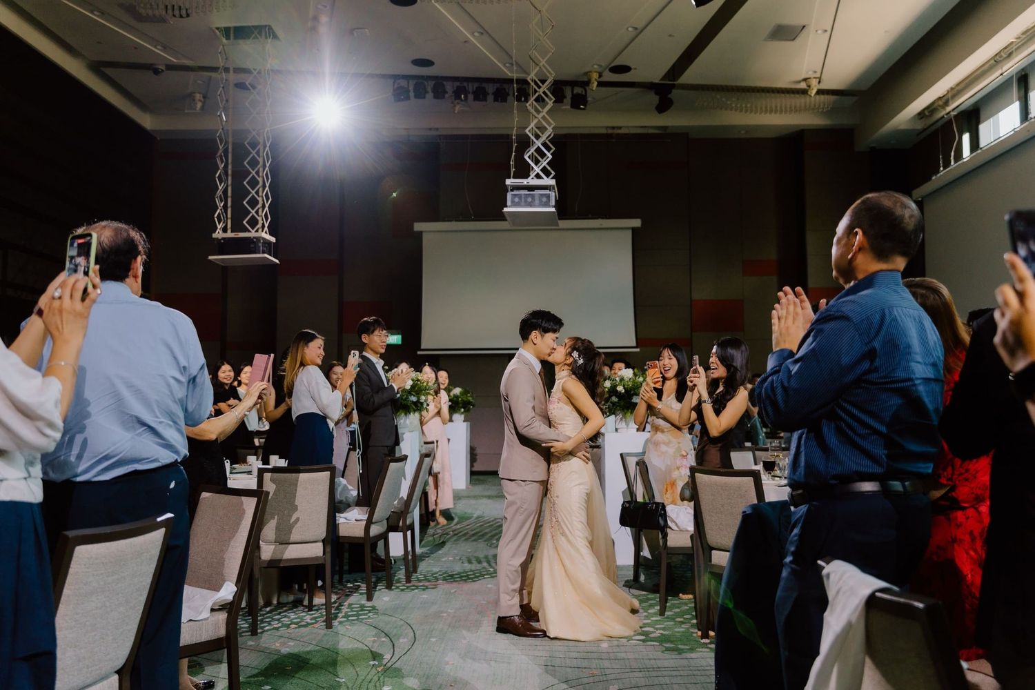 Bride and groom share a kiss as wedding guests applaud in an elegant banquet hall reception venue.