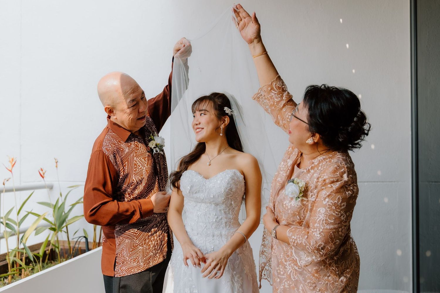 Bride in white gown with parents lifting veil during traditional wedding ceremony blessing.
