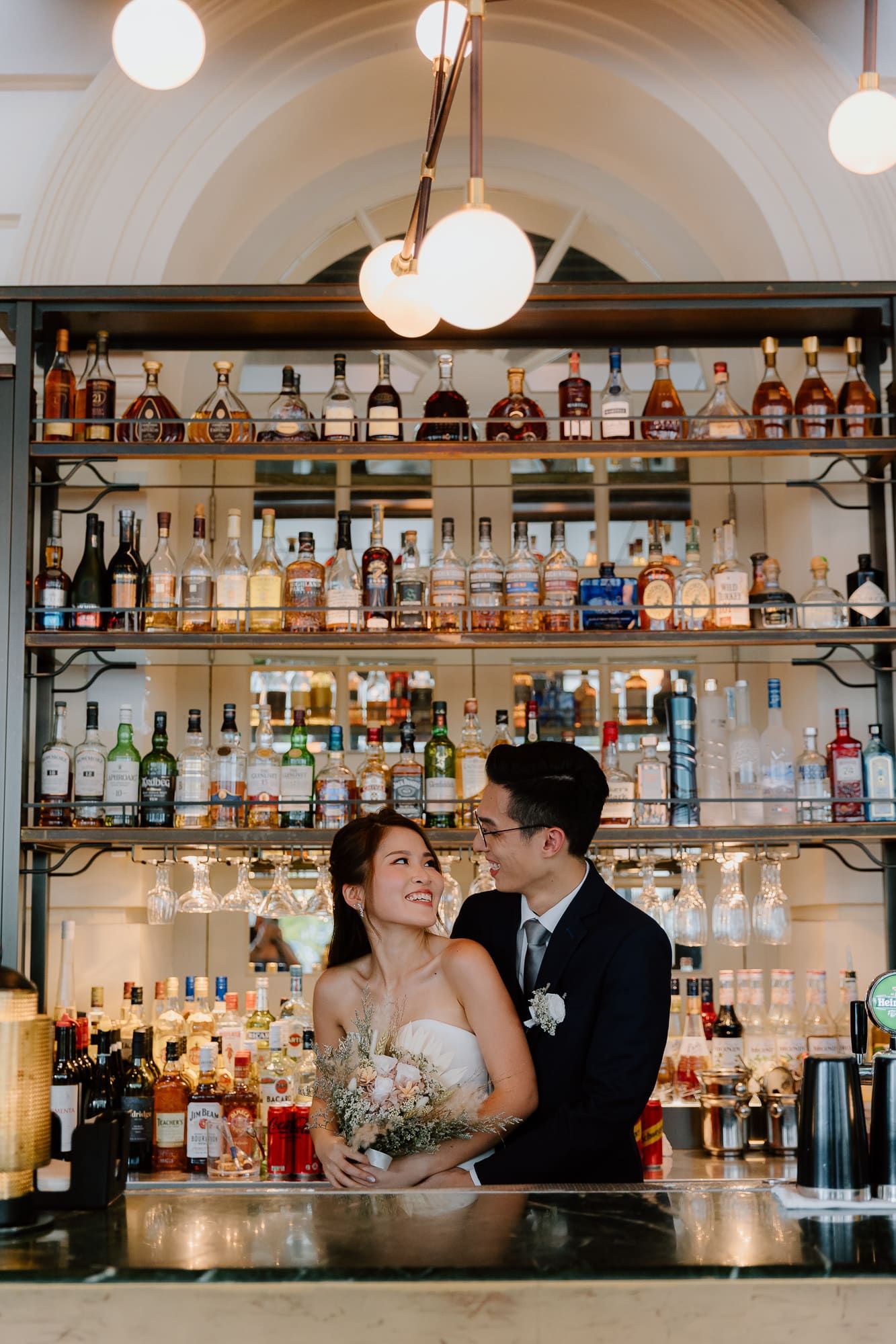 Couple poses together at elegant bar with illuminated globe lights and extensive liquor bottle display on shelves.