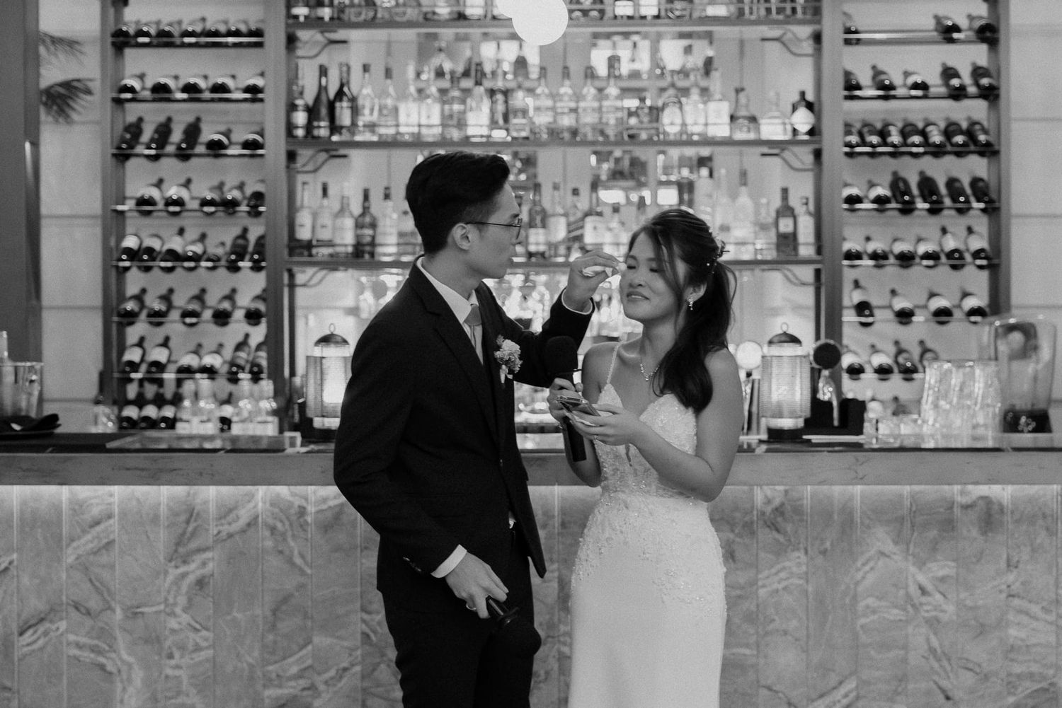 A couple shares a romantic moment at an elegant bar with illuminated shelves of bottles and glassware in black and white.