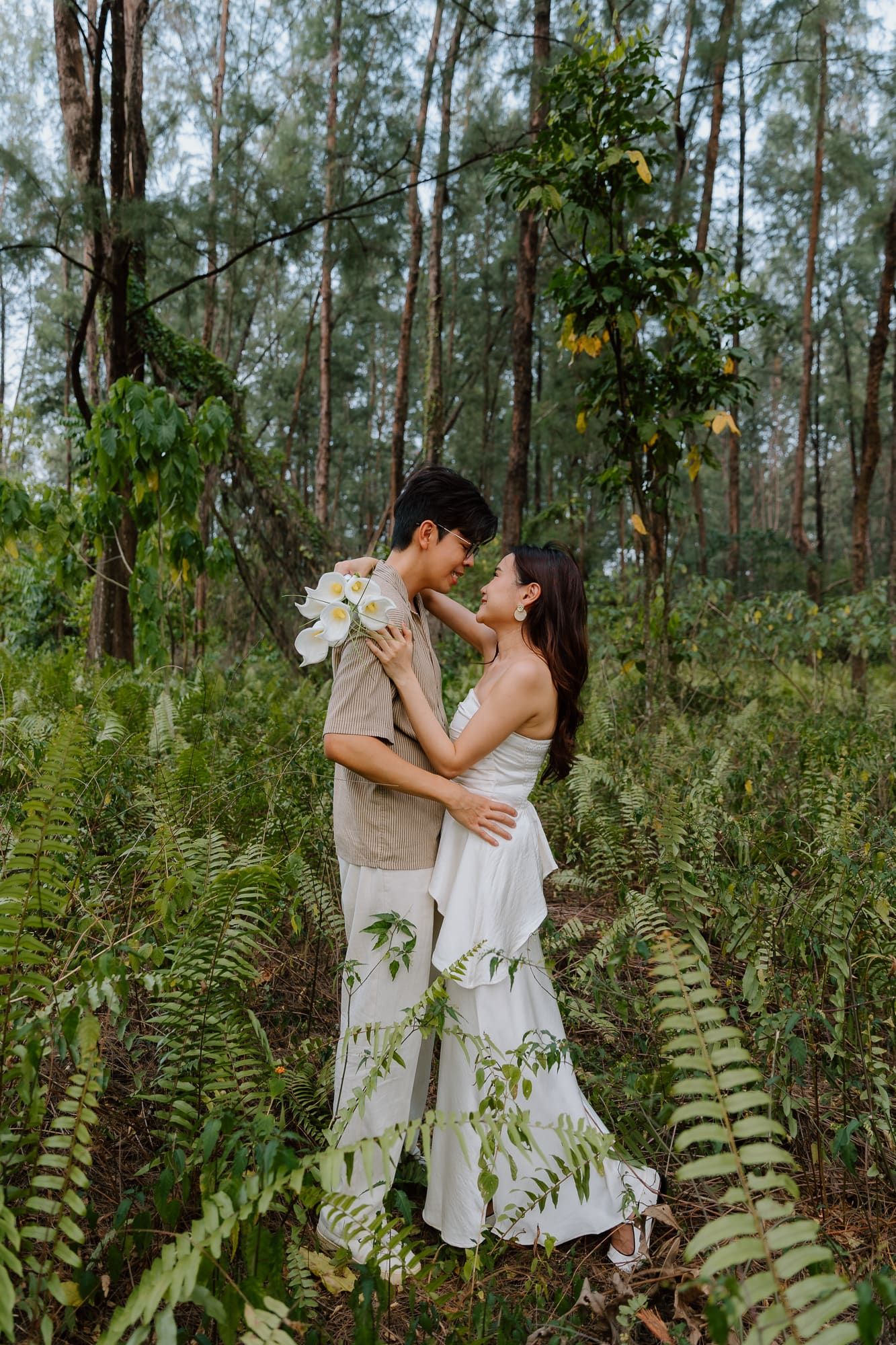 Couple in white dress and beige shirt embracing among ferns in a lush forest setting.