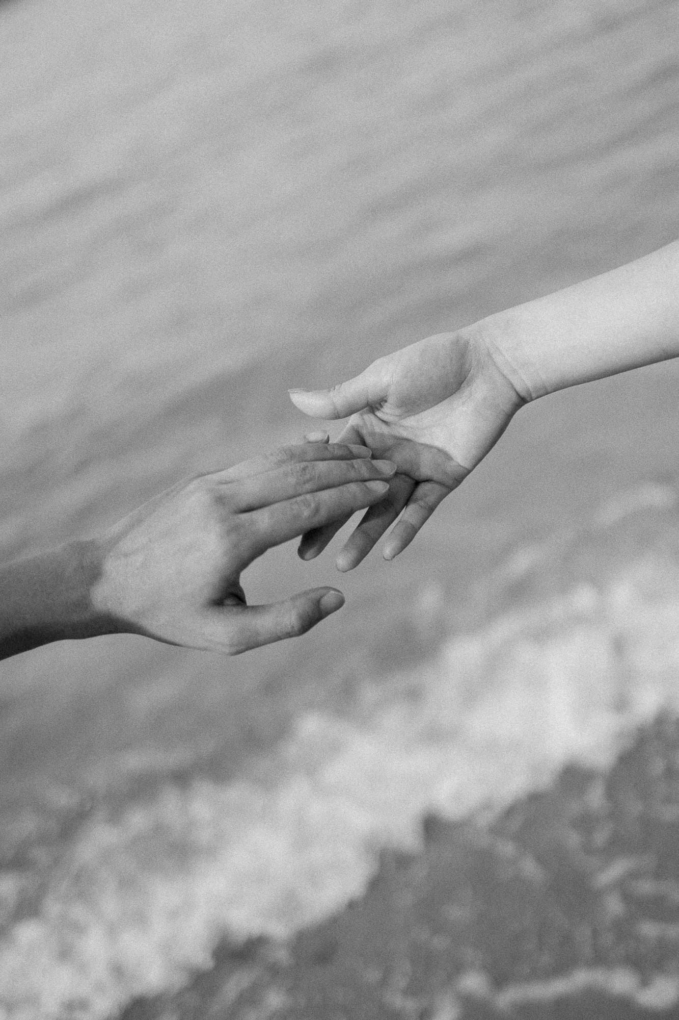Black and white photo of two hands gently touching fingertips near ocean waves, symbolizing love and connection.