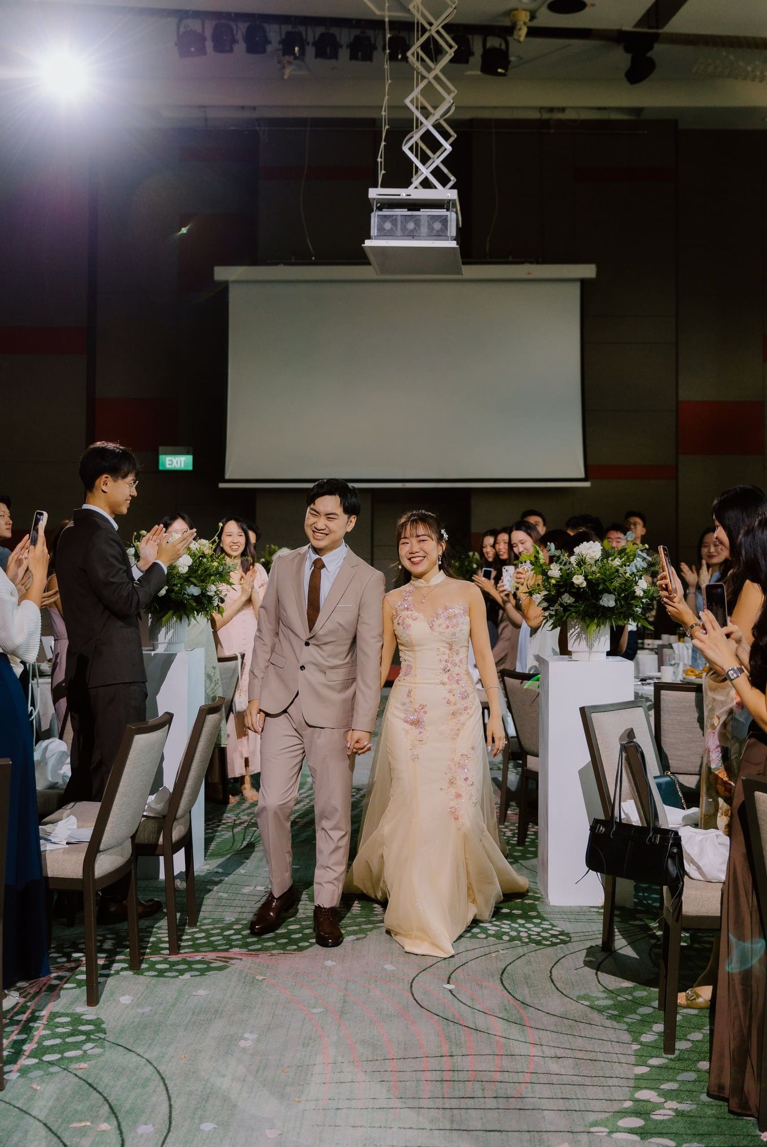 Bride and groom walk down the aisle at an elegant indoor wedding reception, surrounded by guests.