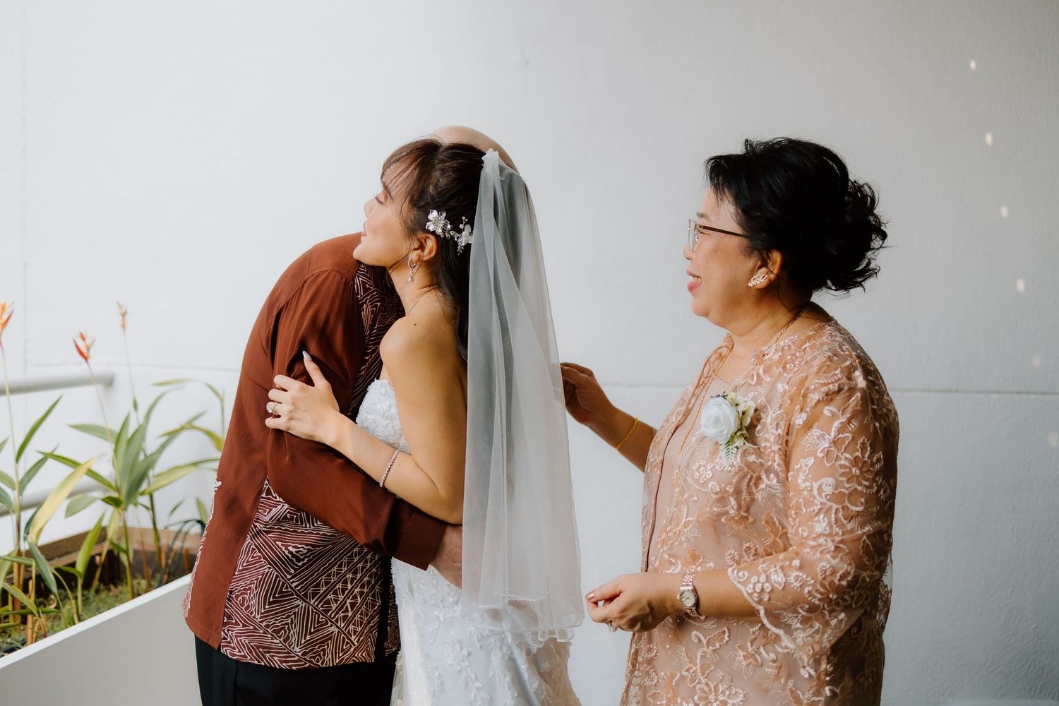 A bride in a white gown and veil receives a kiss on the cheek from a woman in brown while another in gold looks on.