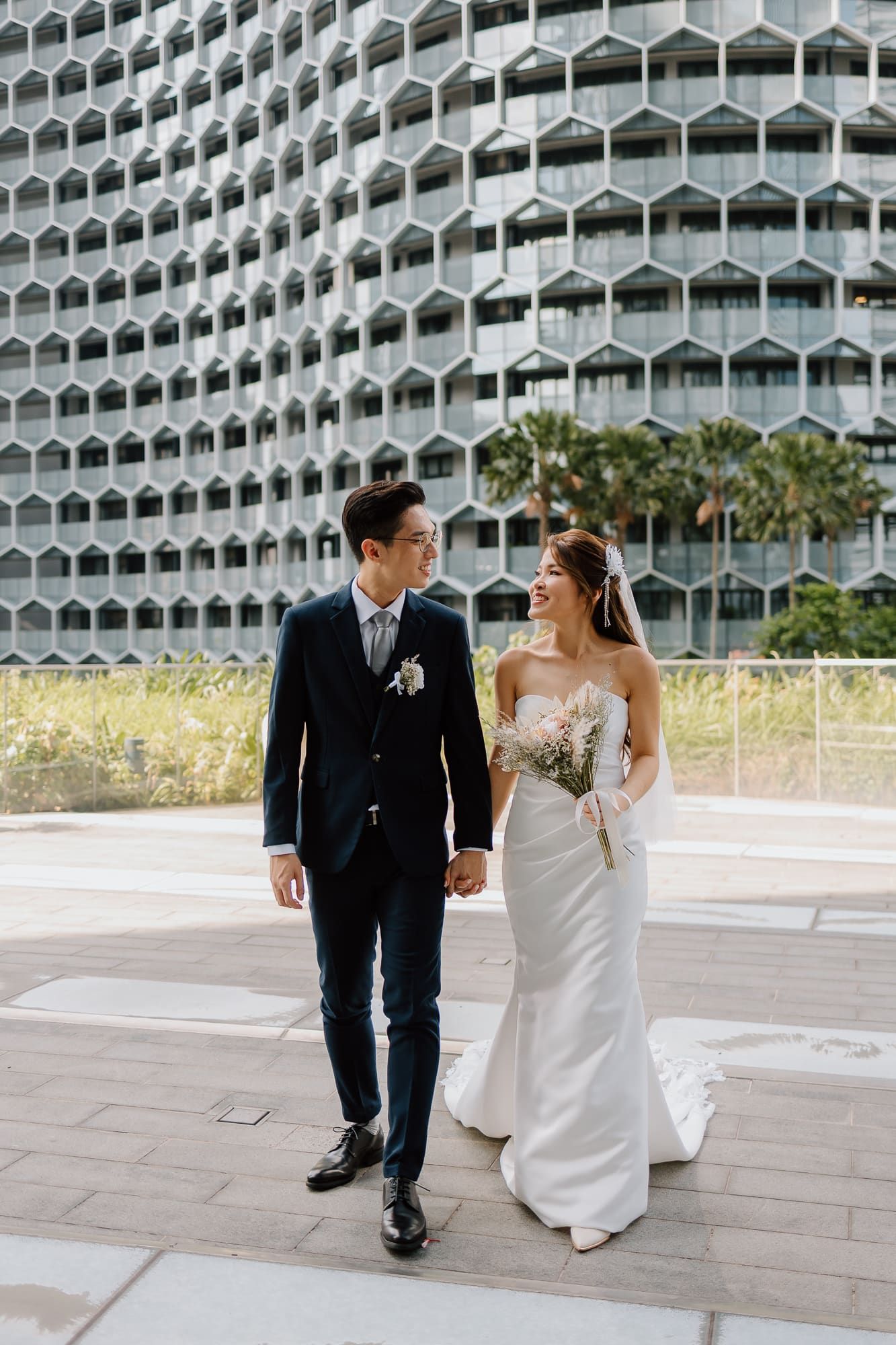 Newlywed couple walks together in front of a modern architectural building with honeycomb-style windows.