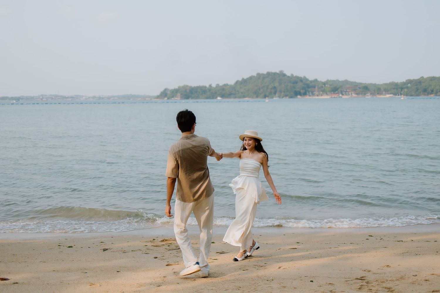 Couple walking hand in hand on a sandy beach, woman in white dress and hat, calm ocean and green island in background.