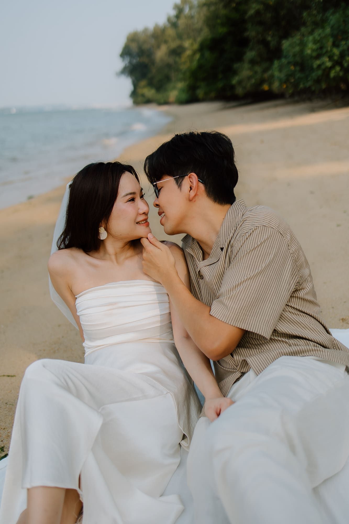 A couple shares a romantic moment on a sandy beach, the bride in a white strapless gown smiling as her partner leans in close.