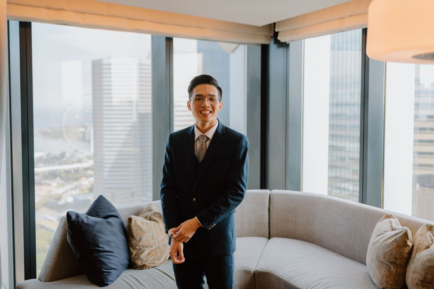 Professional standing by a modern gray sofa in a high-rise office with city views through floor-to-ceiling windows.