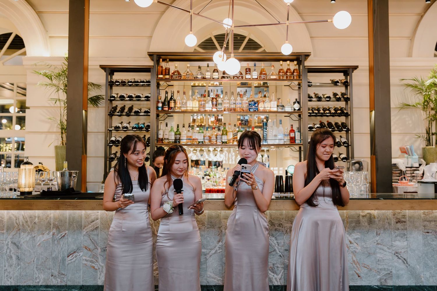 Four people in matching pink dresses stand at an elegant bar counter while checking their phones.