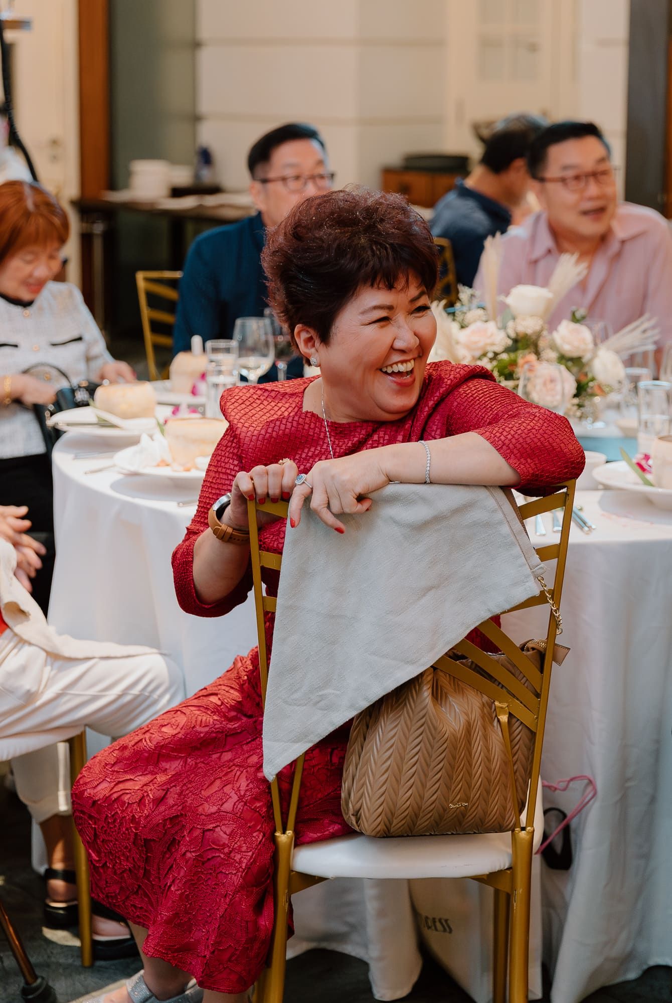 Guest in red lace dress laughs joyfully while sitting at elegant reception table with white linens and floral centerpieces.